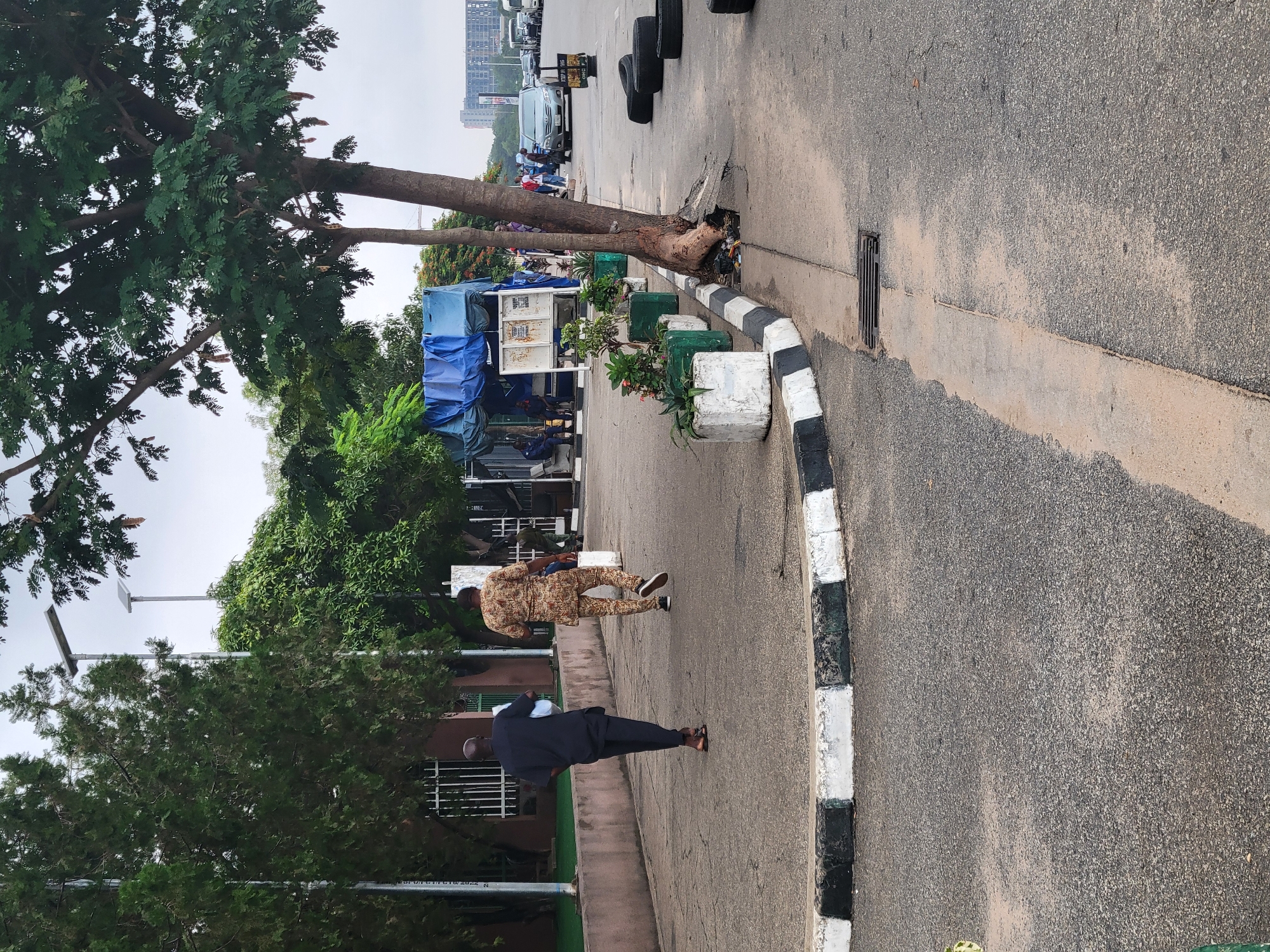 The image depicts a street scene in Abuja, Nigeria, likely in a commercial or administrative area. Two men are walking away from the camera on a paved sidewalk. The man in the foreground wears a black traditional African garment and appears to be carrying a light-colored object. The man behind him is dressed in a patterned traditional African outfit. In the background, a large tree with exposed roots grows adjacent to the sidewalk, which is bordered by a concrete curb with black and white markings. Beyond the curb, a road is visible with parked cars, including what appears to be a silver SUV. A temporary structure covered with a blue tarp is situated further down the street, with people gathered around it. Several large tires are stacked on the side of the road. The sky is overcast. Buildings and trees line the street, and in the far distance, taller buildings suggest an urban environment.