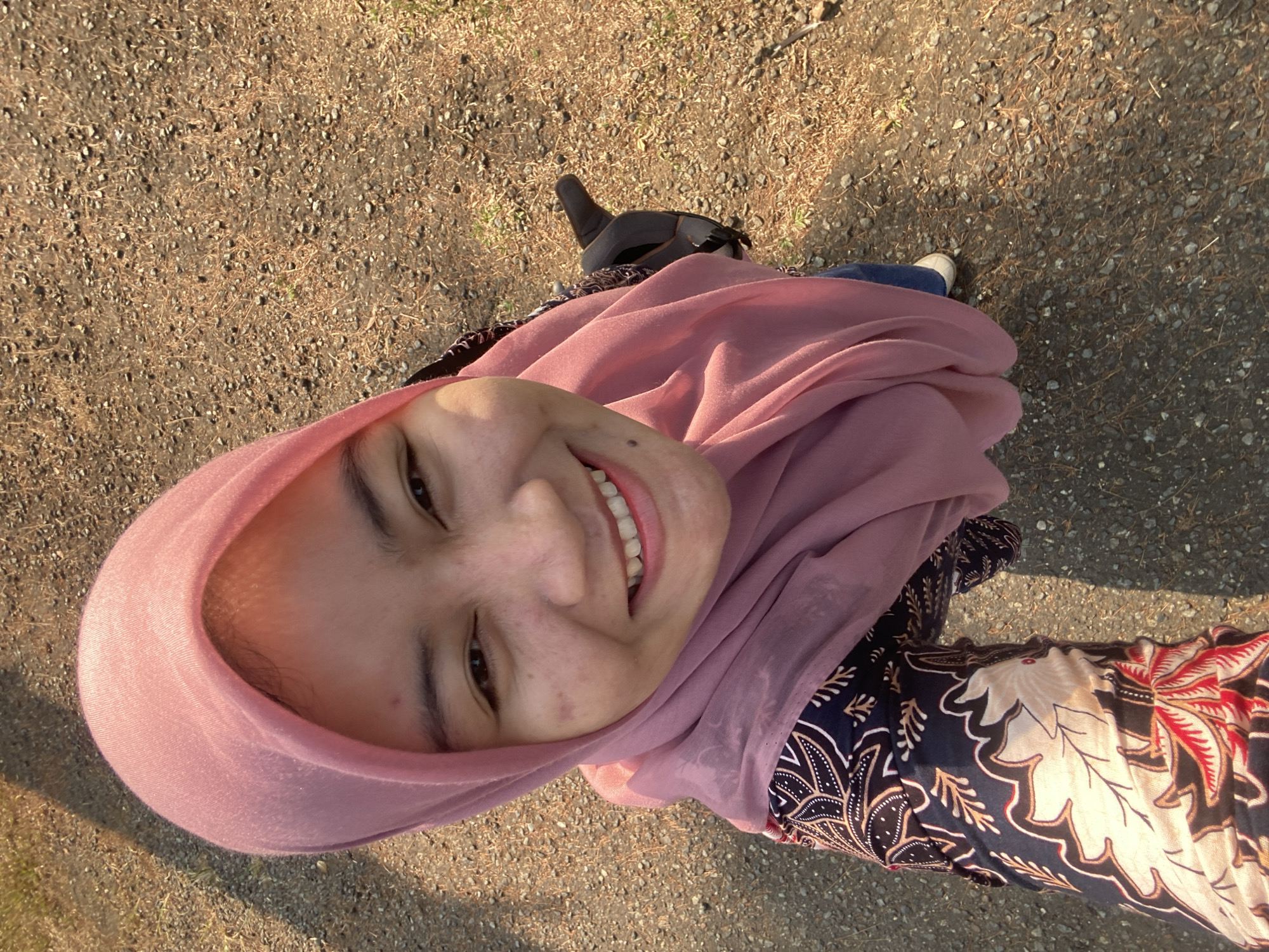 A young woman wearing a pink hijab and a batik-patterned shirt is captured in a high-angle selfie. Her face is angled towards the camera, and she is smiling broadly, revealing her teeth. She has dark eyes and fair skin with some minor blemishes. She is wearing blue jeans and white shoes. Her left arm is visible, holding a black object, possibly a helmet. The background consists of a gravelly path with dry leaves and sparse vegetation, suggesting an outdoor setting. The lighting indicates it is daytime, with sunlight casting shadows. The scene appears to be in a park or a similar outdoor recreational area in Banjarbaru, Indonesia, possibly near a natural setting like a forest edge.