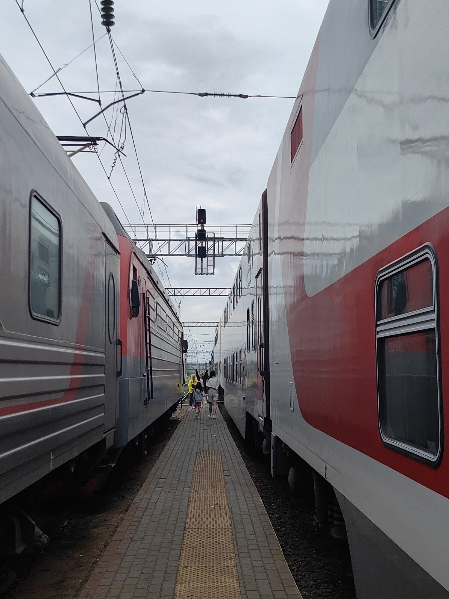 The image captures a railway station scene in Rossosh, Russia. Two trains, one grey and red on the left and the other white and red on the right, stand parallel to each other at the platform. A narrow platform separates the trains. A small group of people, including a child in a yellow jacket, are walking along the platform towards the end of the trains. Overhead, railway infrastructure, including wires and a signal light, are visible against an overcast sky. The platform is paved with brick, and a yellow tactile strip is present.