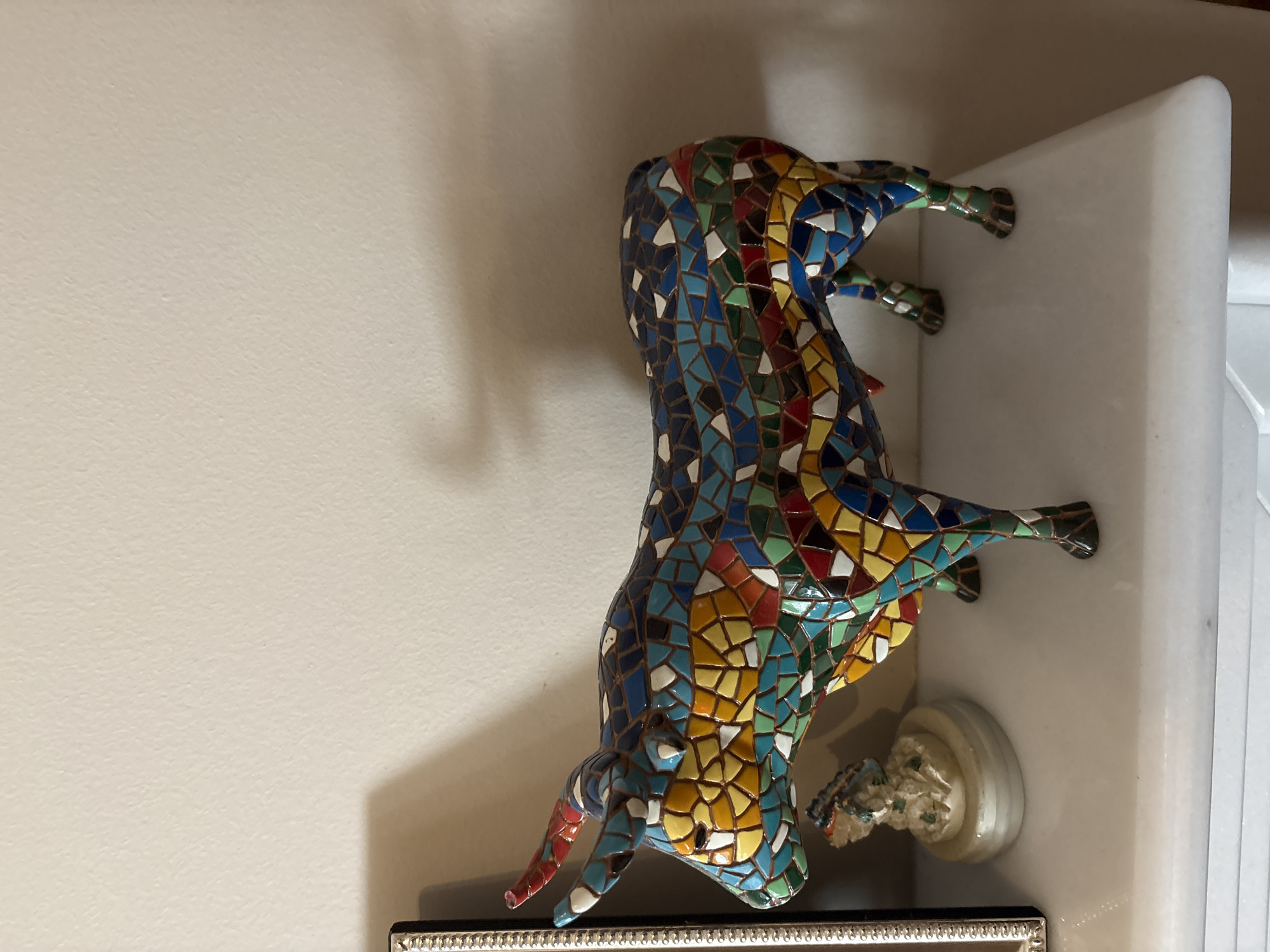 A mosaic bull figurine is displayed on a white shelf against a light-colored wall. The bull is intricately decorated with small, colorful tiles in shades of blue, yellow, red, green, and white, forming a vibrant mosaic pattern across its body. It is positioned with its hind legs resting on the shelf and its front legs extended as if walking or leaping. The bull's head is lowered, and its horns are visible. To the right of the bull, a small, white figurine, possibly a cherub or statue fragment, is also present on the shelf. The shelf itself appears to be made of a white, solid material, possibly marble, with a rounded edge. The background is a plain, textured wall, with a shadow cast on the right side, suggesting indoor lighting. The scene is static, with no apparent activity taking place beyond the display of the objects. The overall impression is one of decorative art within an interior setting. No visible text is present in the image.