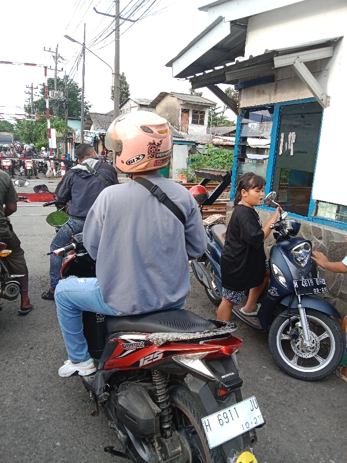A man wearing a pink helmet sits on a red motorcycle with a license plate reading "H 6911 JU," facing right. The man is wearing blue jeans and a grey sweatshirt. The motorcycle is in the foreground, with the handlebars facing right. Another man in a blue shirt and a grey jacket sits on a black motorcycle in the background. Both men are facing left. A young woman with long dark hair wearing a blue sleeveless shirt, white shorts, and black sandals stands next to a blue scooter with a license plate reading "B 4619 BHD." The scooter is facing right. The woman is facing left, looking at a man in a white shirt standing on a white concrete sidewalk. The scene appears to be a typical street intersection in Tugu, Indonesia (latitude: -6.975, longitude: 110.325), with some cars and people in the distance. Buildings on the right side of the image are white with blue windows and doors. The sidewalk in front of the buildings has a concrete border and is lined with small trees. The intersection is a busy area with many motorcycles and cars traveling.