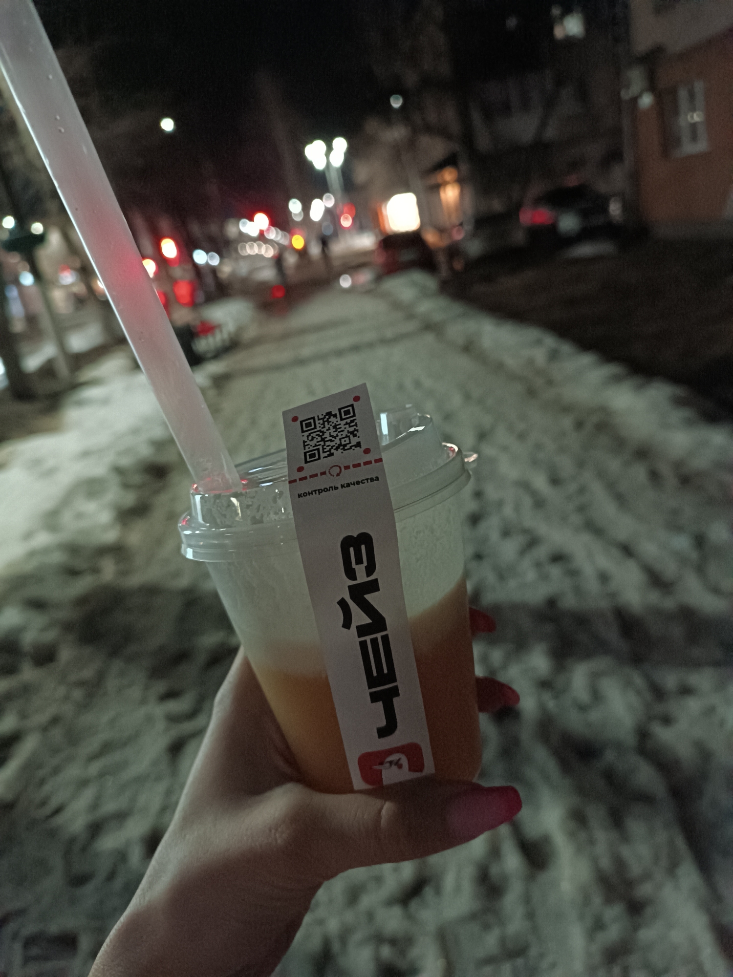 The scene depicts a nighttime outdoor urban environment, specifically a snowy sidewalk. In the foreground, a human hand with pink manicured fingernails holds a clear plastic cup containing a layered beverage, appearing light orange/yellow at the bottom and white/frothy at the top. A translucent white straw extends from the cup's lid. A white sticker is attached to the cup, displaying a QR code at the top, followed by the Cyrillic text "КОНТРОЛЬ КАЧЕСТВА" (Quality Control) and larger Cyrillic text "ЧЕЙЗ" (Chase/Chace), with a small red logo beneath it. The background is out of focus, showing patches of dirty snow on the ground, blurred streetlights, red vehicle taillights, and the indistinct outlines of buildings, including one with an orange-brown facade, suggesting a street in the city of Unknown, Unknown.