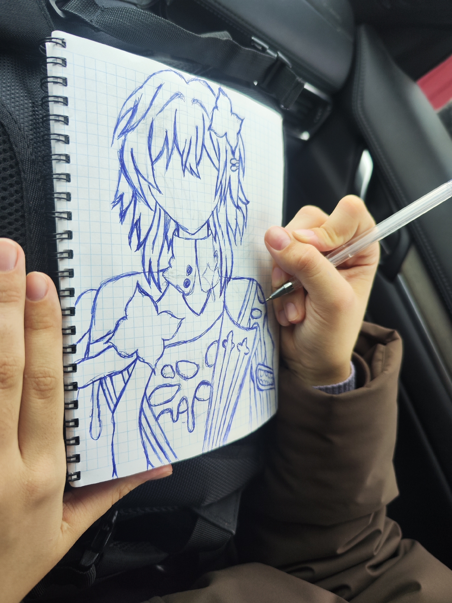 A medium shot captures a person drawing in a notebook. The notebook, featuring a grid pattern and a spiral binding on the left, contains a sketch of a person with stylized hair and intricate clothing details. The sketch is drawn in blue ink. The person drawing is holding a transparent pen in their right hand, poised to add more details to the sketch. Their left hand is holding the notebook open.
The background is slightly out of focus, but suggests the interior of a car. The seat is black leather, and a part of a seatbelt is visible at the top of the frame. The person drawing is wearing a brown jacket with a hint of a gray sweater underneath.
The location, Chaikovsky, Russia, cannot be verified from the visual information in the image.