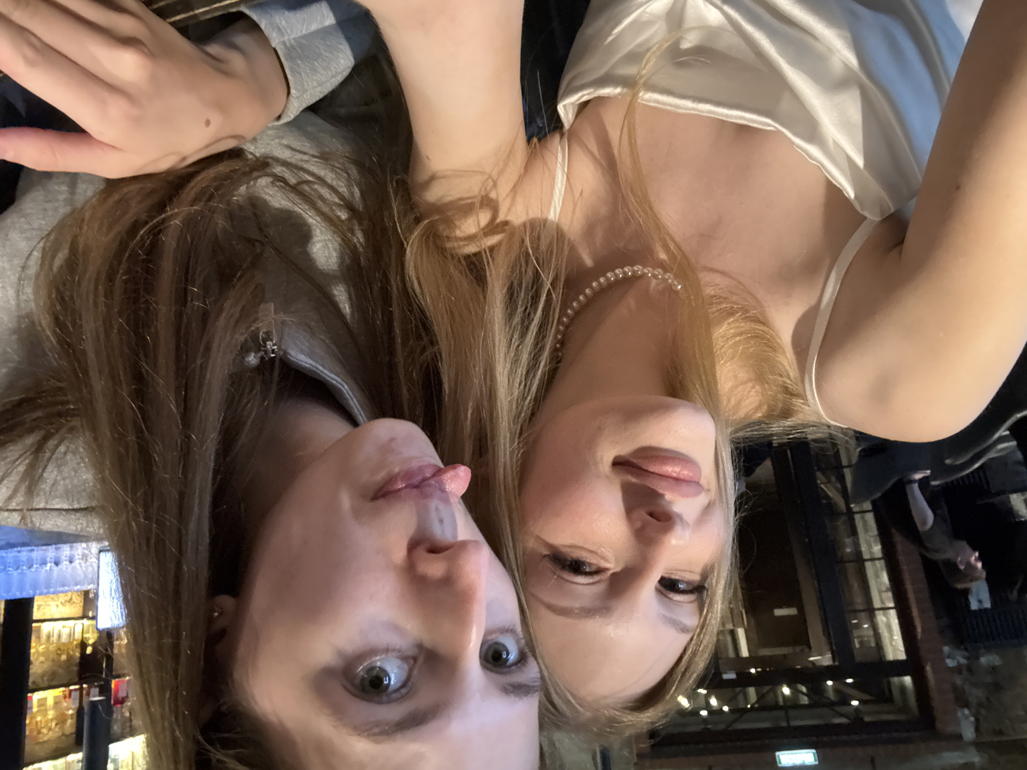 Two young women are taking a selfie in what appears to be a dimly lit bar or restaurant. The woman on the left is smiling and wearing a white satin slip dress with a pearl necklace. The woman on the right is sticking out her tongue and wearing a grey hooded jacket. In the background, there are shelves stocked with bottles, suggesting a bar area. A man in a black t-shirt is visible in the far left background, walking away from the camera. The overall atmosphere is casual and social. This location is not identifiable as being in Perm, Russia, or any specific city.