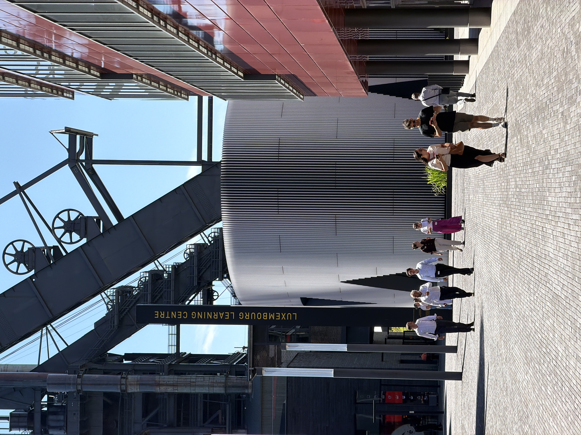 The image depicts the industrial heritage site of Belval in Esch-sur-Alzette, Luxembourg. In the foreground, a paved plaza is populated by several people walking. From left to right, there are three men in business attire walking together, followed by a couple walking and looking at their phones, and a man in casual wear walking away from the camera. To the right, a modern red building with reflective windows is visible. In the background, the imposing industrial structures of the former steelworks dominate the scene, including large metal beams and machinery, juxtaposed with a sleek, cylindrical modern building, likely the Luxembourg Learning Centre. A tall, black sign with yellow lettering reads "LUXEMBOURG LEARNING CENTRE". The sky is clear and blue, indicating a sunny day.