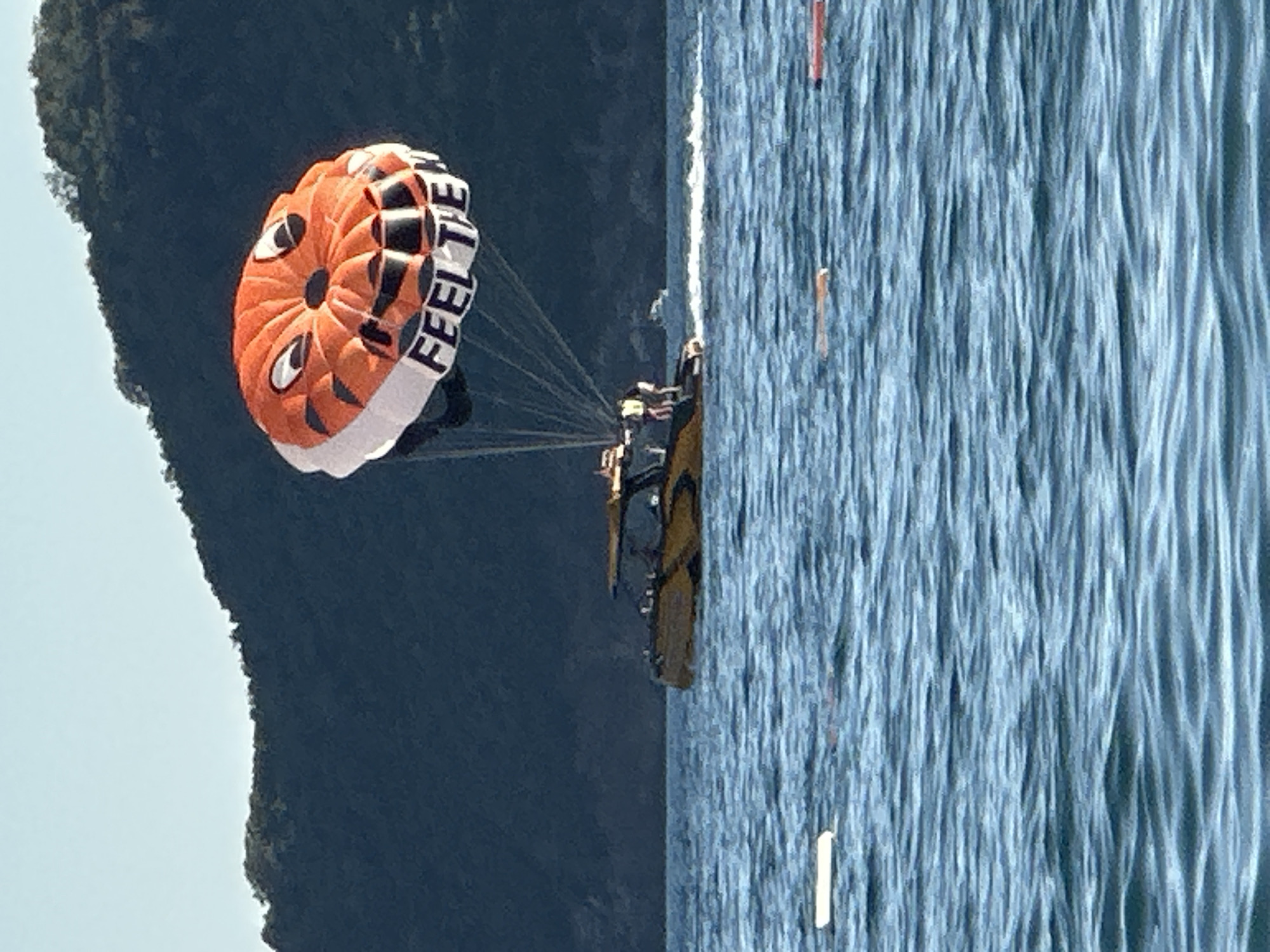 In the waters off the coast of Boreti, Montenegro, a boat is seen with several people on its deck. The boat is moving, creating a wake. Above the boat, a large, orange and white parachute is inflated and in the air, attached to the boat by ropes. The parachute is decorated with a cartoonish tiger face and the words "FEEL THE". The water is blue with moderate ripples. In the background, a dark, tree-covered hill rises against a pale blue sky. Several buoys are visible floating on the surface of the water.