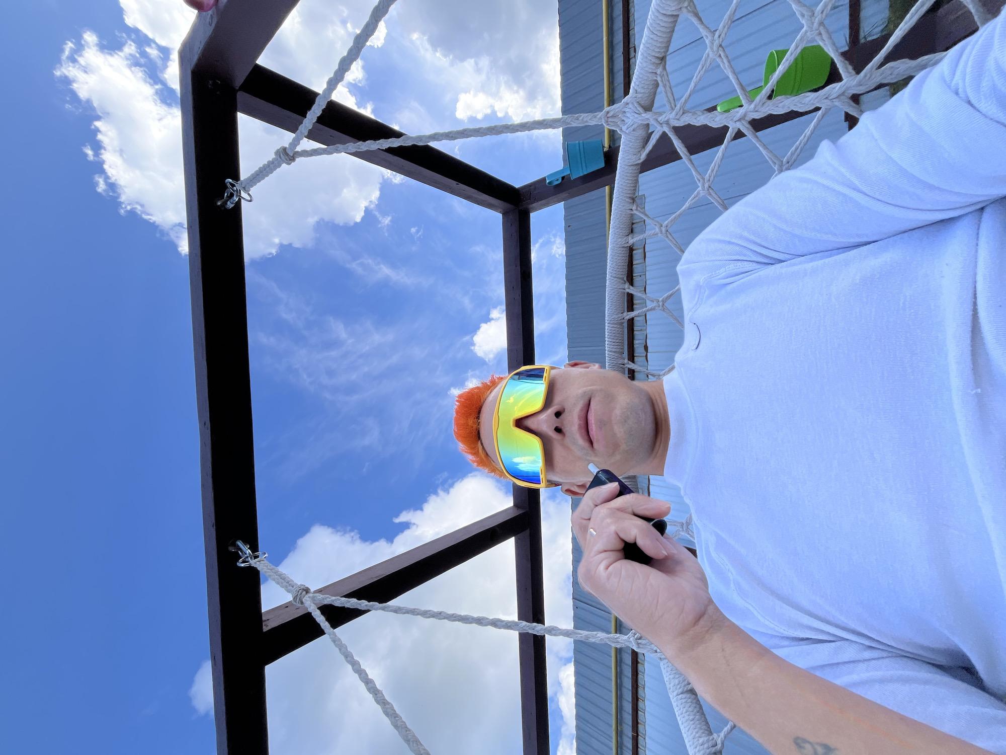 A man with orange hair and iridescent sunglasses, wearing a white T-shirt, is holding a small black object in his right hand. He's in a hammock suspended from a dark-colored wooden frame. The sky, filled with white clouds, is visible through the frame. The side of a building with corrugated metal is also visible. The image was taken in Chaikovsky, Russia.