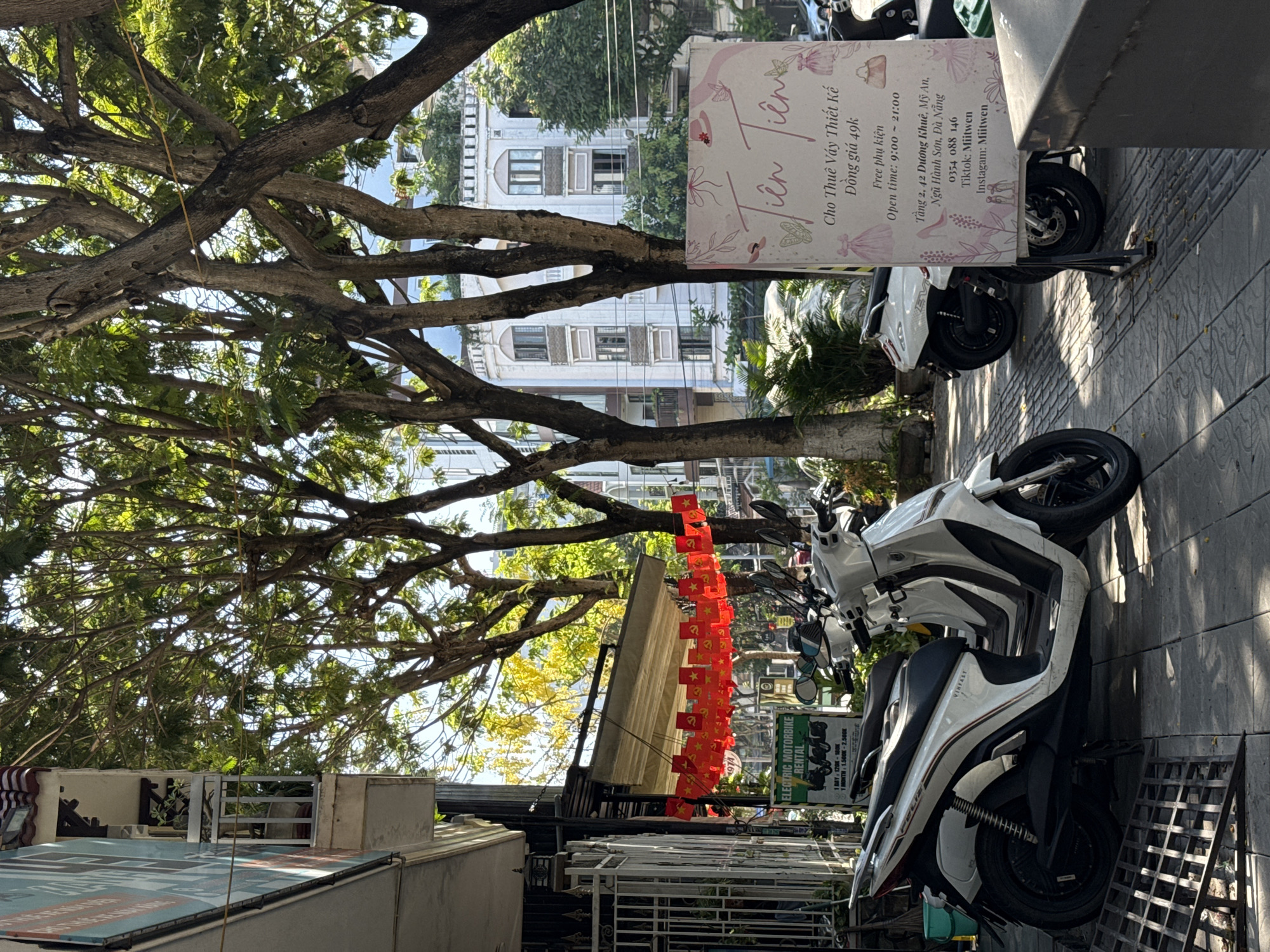 The image shows a street scene in Đà Nẵng, Vietnam. Several white VinFast electric motorbikes are parked on the sidewalk. A sign advertises "Tiên Liên" dress rentals for 49k đồng, located at 42 Dương Khuê, Mỹ An, Ngũ Hành Sơn. The sign also promotes the business's TikTok and Instagram accounts, Miitwen. Above, a line of small red Vietnamese flags hang between buildings. A large tree with dense foliage dominates the upper left corner, partially obscuring a white building in the background.