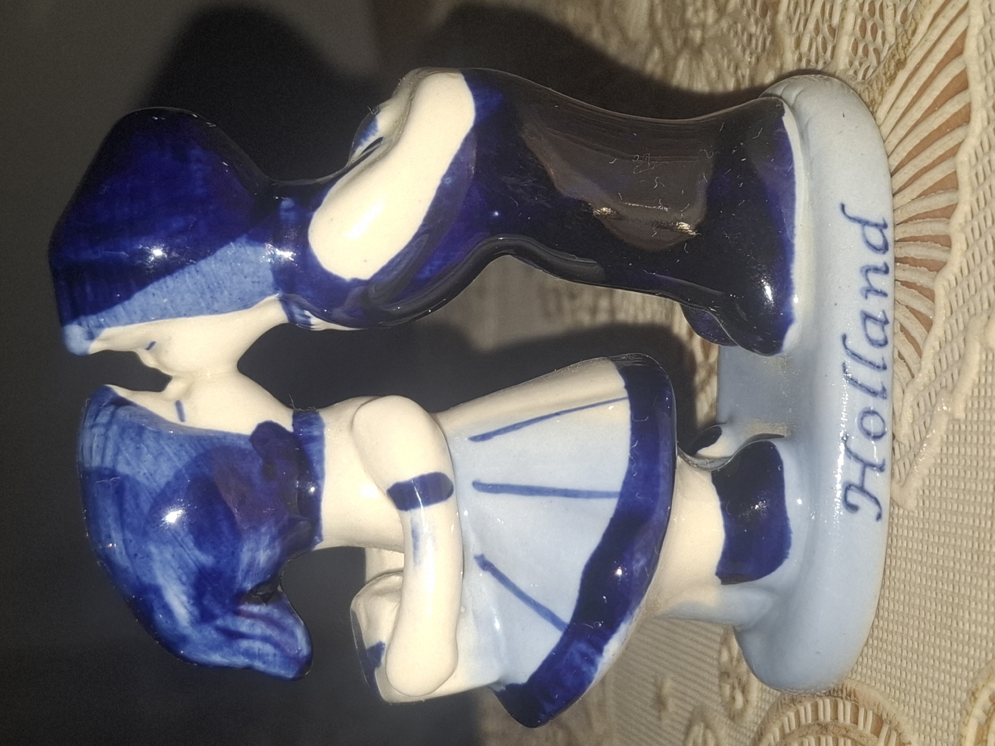 A ceramic figurine depicts two stylized figures, possibly children, kissing. The figures are predominantly white with blue accents, painted in a Delftware style. The left figure wears a blue bonnet and a white dress with blue stripes and trim. The right figure wears a blue hooded garment. They are standing on a light blue base inscribed with the word "Holland" in blue cursive lettering. The figurine is placed on a textured surface, possibly a tablecloth, with a dark background behind it. There is no indication of a city, location, or any specific environment beyond the immediate placement of the object.