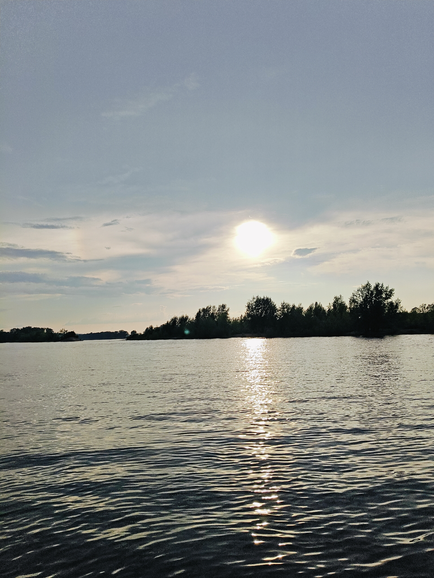 The image depicts a wide, calm river flowing through a natural landscape under a partly cloudy sky. The sun is visible, high and bright, with its reflection creating a shimmering path on the water's surface. On the far bank of the river, a dense line of trees forms a dark silhouette against the sky. The foreground is dominated by the rippling surface of the river, with distinct patterns of light and shadow created by the sun. The overall atmosphere is serene and tranquil, suggesting an outdoor scene in a natural setting. The location is identified as Noviy, Russia.