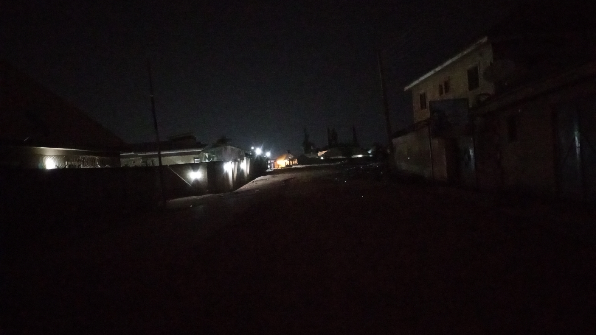 This media file captures a dark, quiet street at night in Sabon Tasha, Nigeria. The scene is illuminated primarily by artificial lights emanating from buildings and wall-mounted fixtures.
The road, appearing unpaved or dirt, slopes gently uphill into the distance. On the left side of the frame, a substantial fence with several bright wall lights runs alongside the road, casting illumination onto the ground and part of the fence itself. Above this fence, what appears to be barbed wire or razor wire is visible, suggesting security measures. Beyond the fence, the silhouette of a building's roofline can be seen.
On the right side of the road, a two-story building with a light-colored exterior and a sloped roof stands out against the dark sky. A satellite dish is mounted on its roof. Adjacent to it, a darker, single-story structure with a doorway is visible. Further down the street, more structures with distant lights punctuate the darkness. Utility poles are present on both sides of the road, extending vertically. In the mid-ground on the right, some debris or litter is scattered on the ground, dimly lit by the distant sources.
The sky is a deep, dark black, indicating deep night. There are no visible clouds or stars, and no natural light sources like the moon are prominent. No people or specific activities are observable in the image, contributing to the quiet, still atmosphere. There is no visible text. The overall impression is that of a residential or mixed-use area relying on localized lighting after sunset.