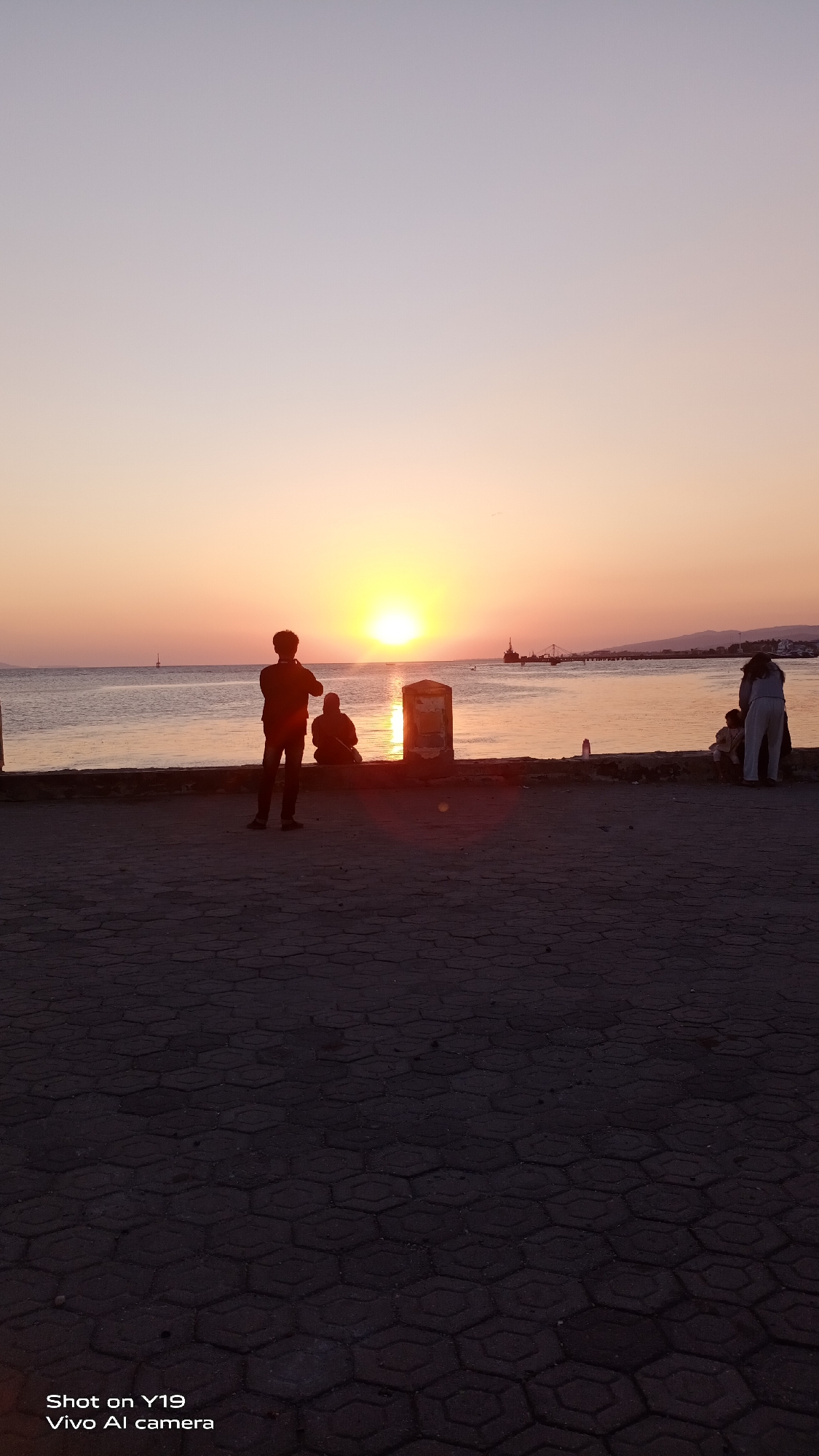 The image depicts a sunset scene by the sea, likely in Sea, Indonesia, given the "Vivo AI camera" watermark. Silhouettes of people are visible along a waterfront promenade. In the foreground, a person stands facing away from the camera, looking towards the setting sun. Further along the promenade, another person is seated. To the right, a group of people, including children, are gathered. The sun is low on the horizon, casting a warm glow across the water, which is calm. A ship or boat is visible in the distance. The ground in the foreground is paved with hexagonal tiles. The sky transitions from a pale orange near the horizon to a soft purple higher up.