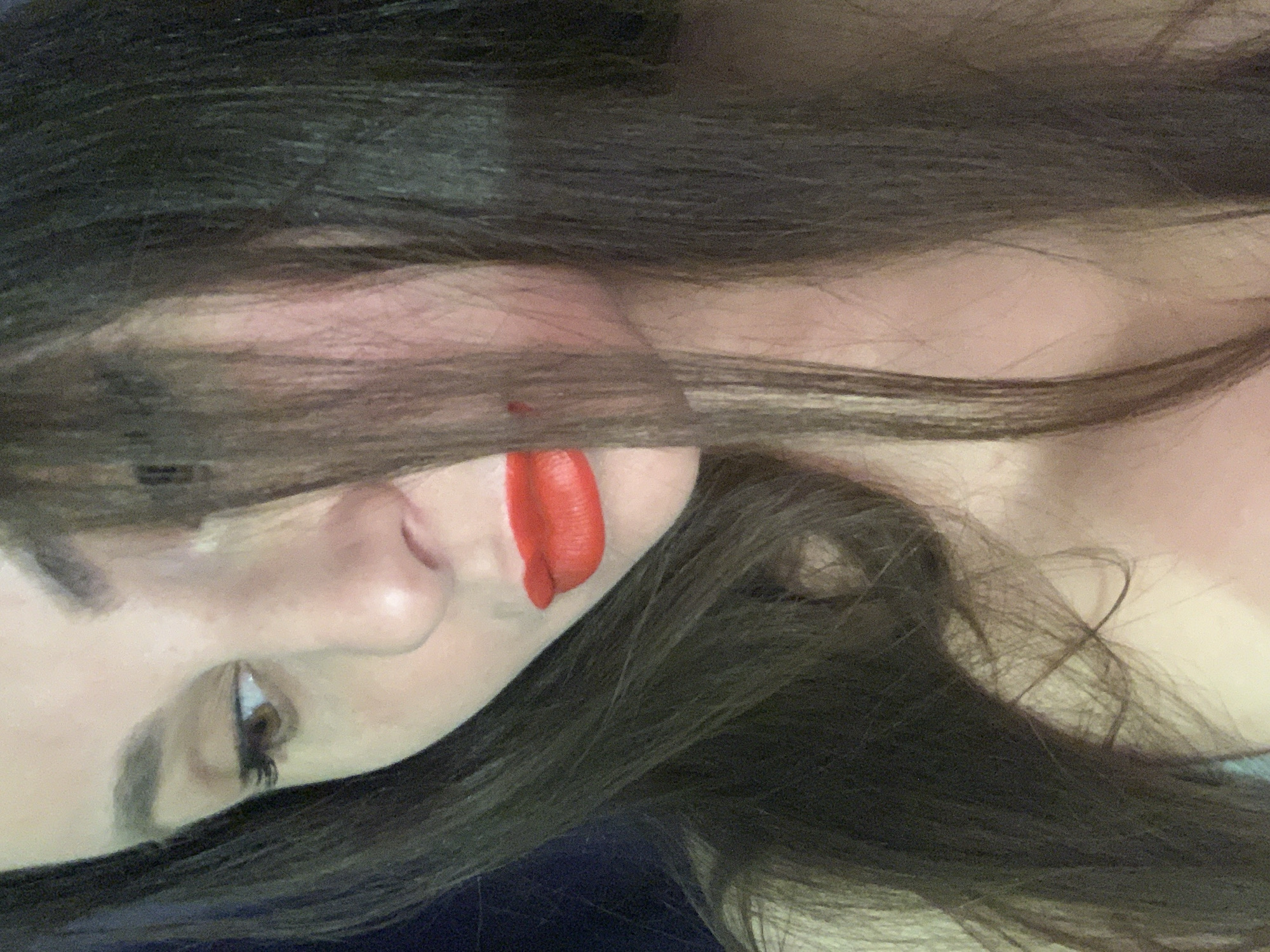 A person with long dark hair is partially visible in a close-up shot. The individual has fair skin, a visible eye with brown lashes, a nose, and lips painted with bright red lipstick. The hair obscures parts of the face. The background is out of focus and dark. The image is rotated, so the person appears sideways. The location is unknown.