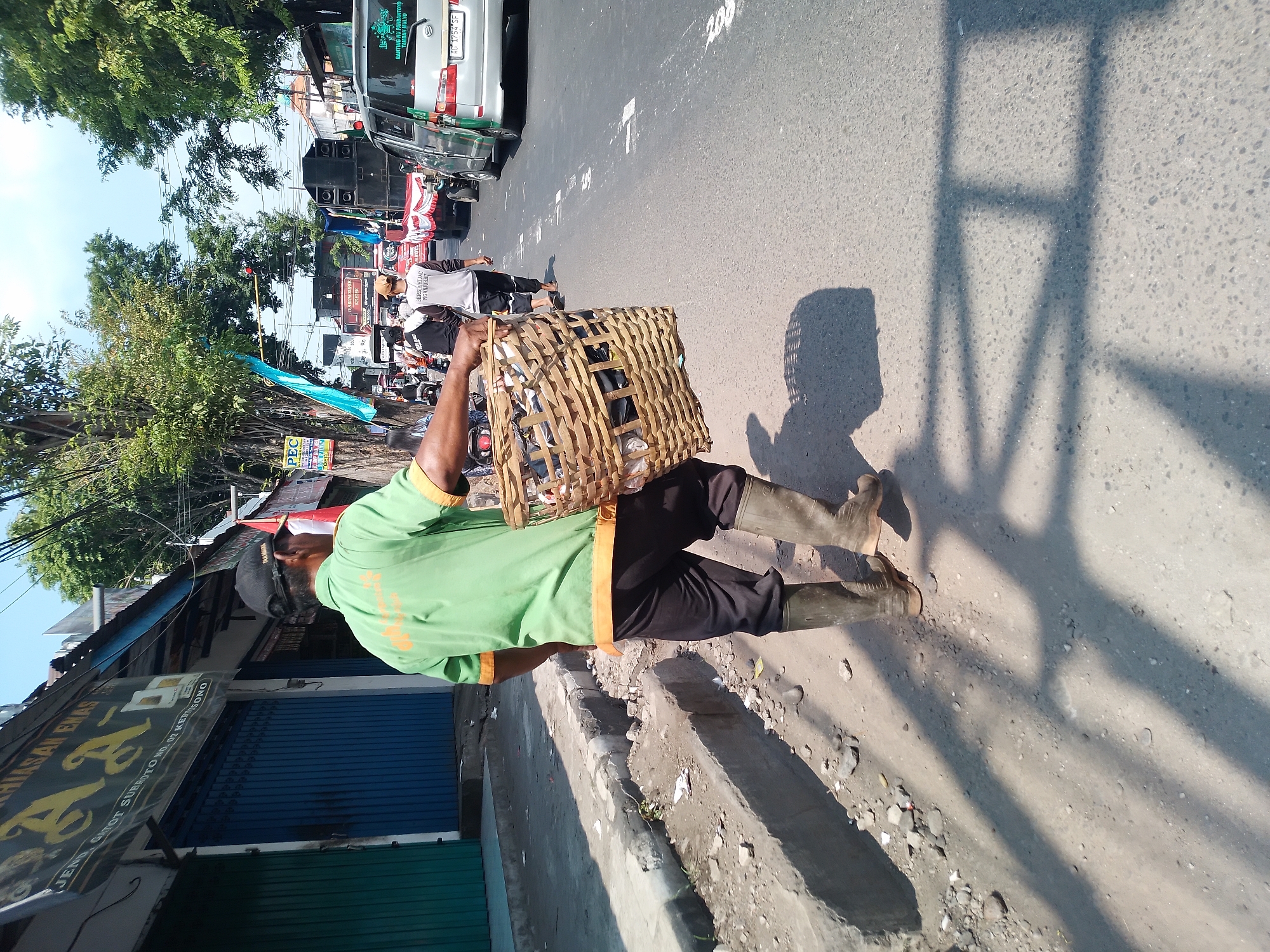 In Kertosono, Indonesia, a man wearing a green shirt, black pants, and tall rubber boots walks on the right side of a paved road, carrying a woven basket filled with items. He is walking away from the camera towards the left side of the frame. To his left is a dirt embankment with a concrete curb, and beyond that are closed storefronts with blue and green metal shutters. The storefronts have signs, one of which appears to say "JENS CNI GHOT SUBITO NO. 02 KERTOSONO". The road is moderately trafficked with cars and motorcycles visible in the background. Trees line the street, and power lines are strung overhead. The scene appears to be a commercial street during daylight hours.