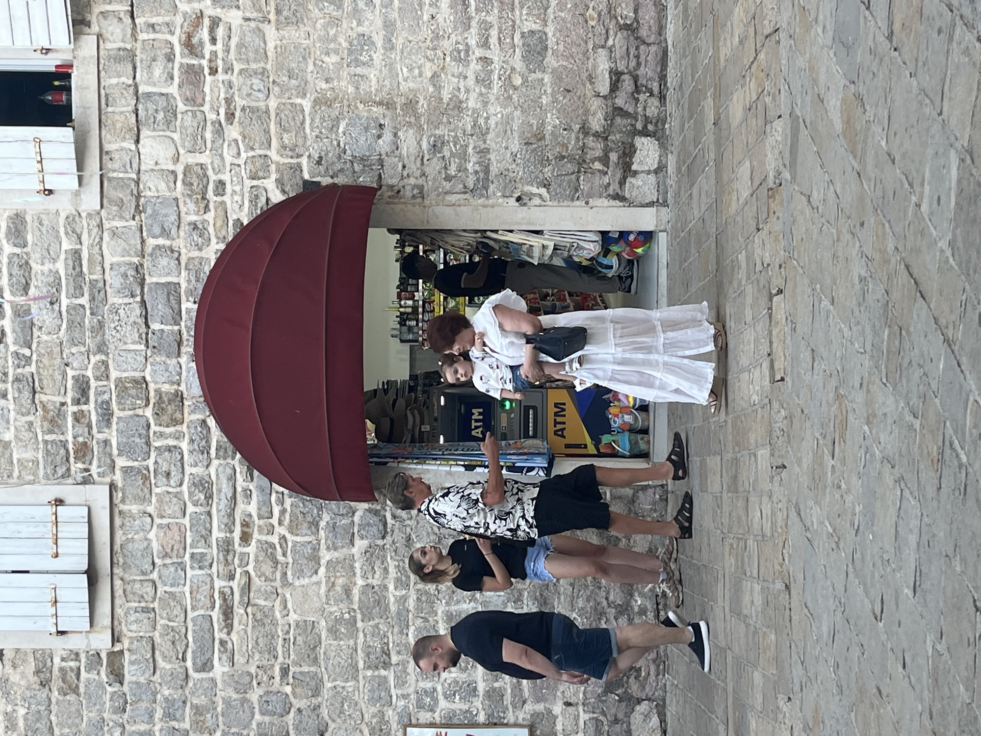 This image depicts a street scene in Budva, Montenegro, near the Old Town. A group of people are gathered outside a shop with a prominent red awning. The shop appears to be a convenience store or souvenir shop, with ATMs visible near the entrance. The architecture features a rustic stone wall.
In the foreground, a man in a black t-shirt and denim shorts is walking away from the camera. To his right, a young woman in a black t-shirt and denim shorts is standing. A man in a patterned short-sleeved shirt and black shorts is speaking to a woman who is holding a young child. The woman is wearing a white dress. The child is wearing a white outfit. Behind them, a man in a black t-shirt and grey pants is inside the shop, possibly an employee.
The ground is paved with cobblestones. In the background, the stone wall of a building is visible, with a window with light blue shutters. The shop's entrance shows shelves stocked with various items, including bottles and what appear to be beach toys. The overall atmosphere suggests a warm, possibly late afternoon, setting.