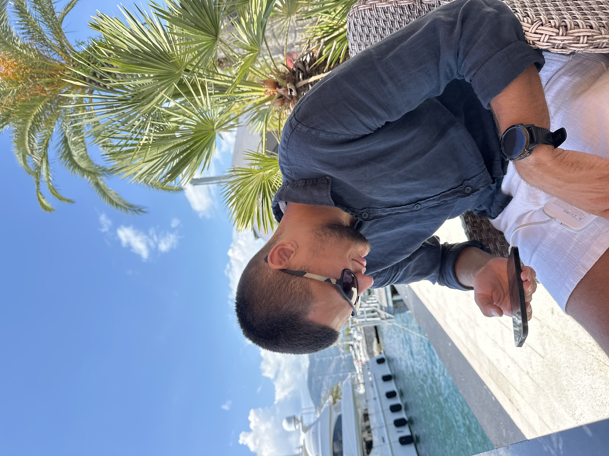 In Tivat, Montenegro, a young man with a short haircut and sunglasses is seated outdoors at a table, looking down at his smartphone. He is wearing a dark linen shirt and white shorts. The background shows a harbor with a large white yacht docked in the turquoise water. Palm trees and a bright blue sky with scattered clouds complete the scene. The man has a watch on his left wrist.