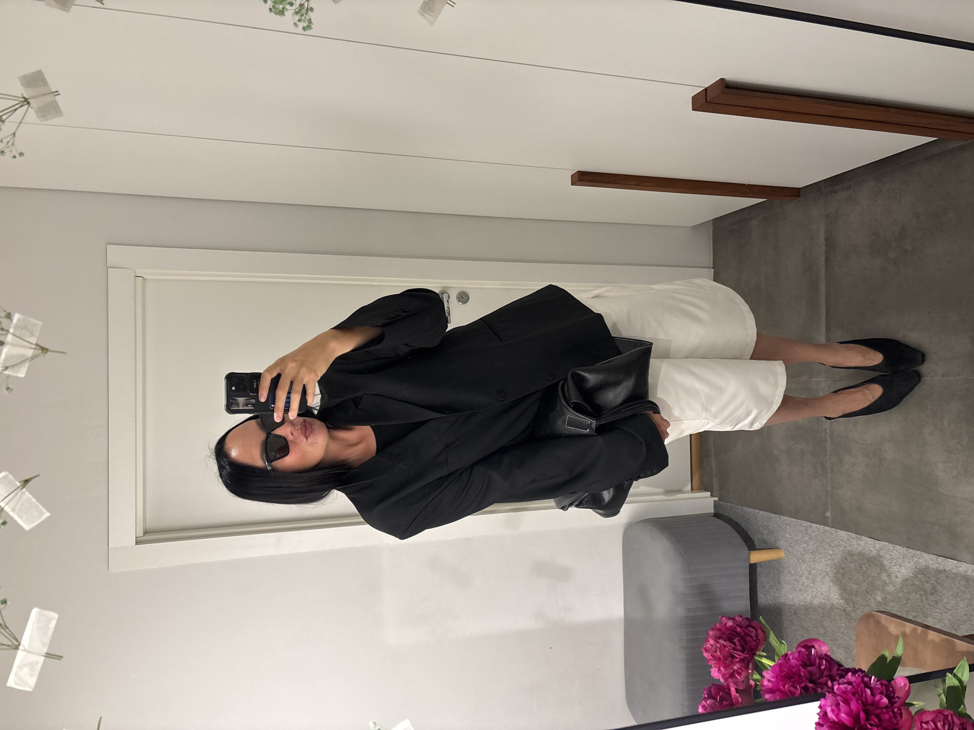A woman is taking a selfie in a brightly lit room with white walls and gray flooring. She is wearing a black blazer, a black top, white shorts, black pointed-toe heels, and black sunglasses. She is holding a black handbag and a phone, and she is puckering her lips. In the background, there is a gray ottoman with wooden legs, a white door, and a full-length mirror that reflects the scene. There are also some pink flowers on the left side of the image. The location is not Chaikovsky, Russia.
