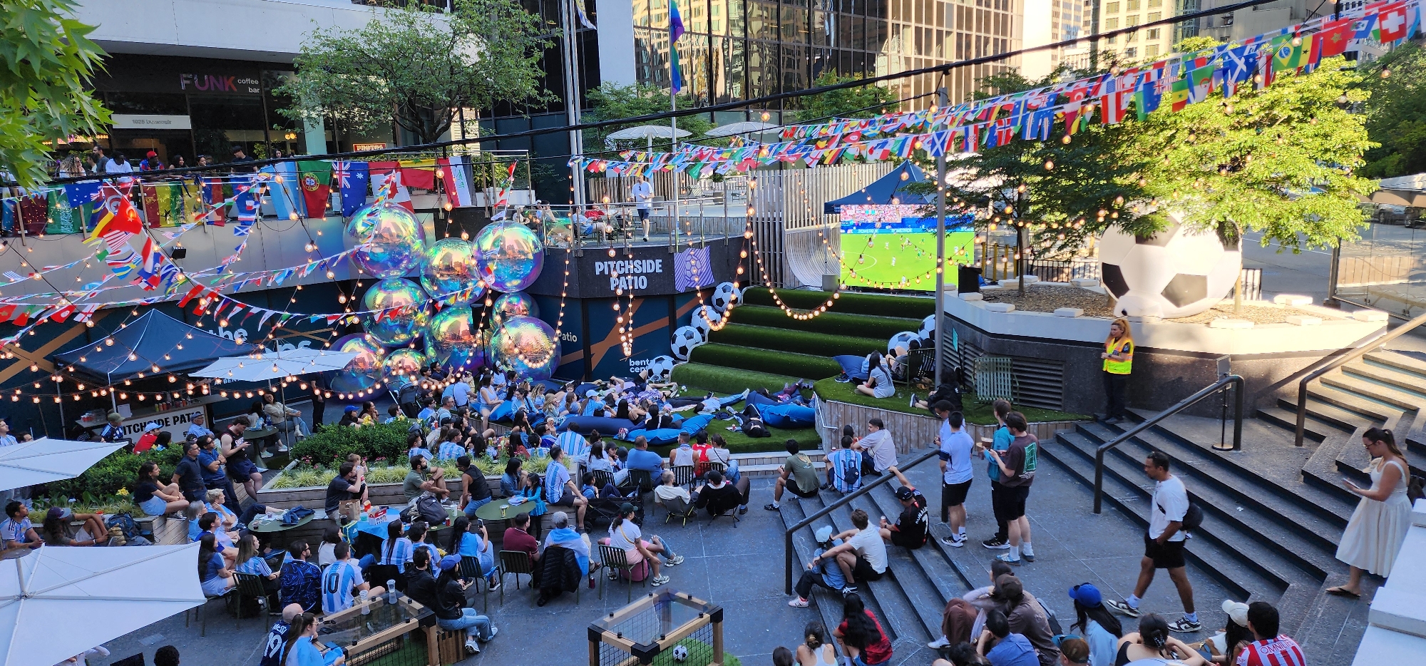 This is an outdoor scene depicting a soccer-themed event, likely a viewing party for a match. The location is an urban setting, with tall buildings visible in the background. The scene is filled with people of various ages, many wearing jerseys, suggesting they are fans.
In the foreground, a large gathering of people are seated on the ground, on beanbags, and in low chairs, facing a large outdoor screen where a soccer game is being projected. The area is decorated with numerous national flags strung together, forming a festive atmosphere. Large, iridescent spherical decorations are also prominent in the scene, adding to the visual spectacle.
Further back, a raised platform labeled "PITCHSIDE PATIO" is visible, with more seating and people watching the screen. A large, inflatable soccer ball is positioned on a raised area to the right, near a set of stairs where other attendees are standing or walking. String lights are draped across the area, illuminating the scene.
The overall environment appears to be a well-decorated urban plaza or courtyard, adapted to host a communal viewing experience for a soccer event. The attendees are casually dressed, and many are engaged in watching the game or socializing.