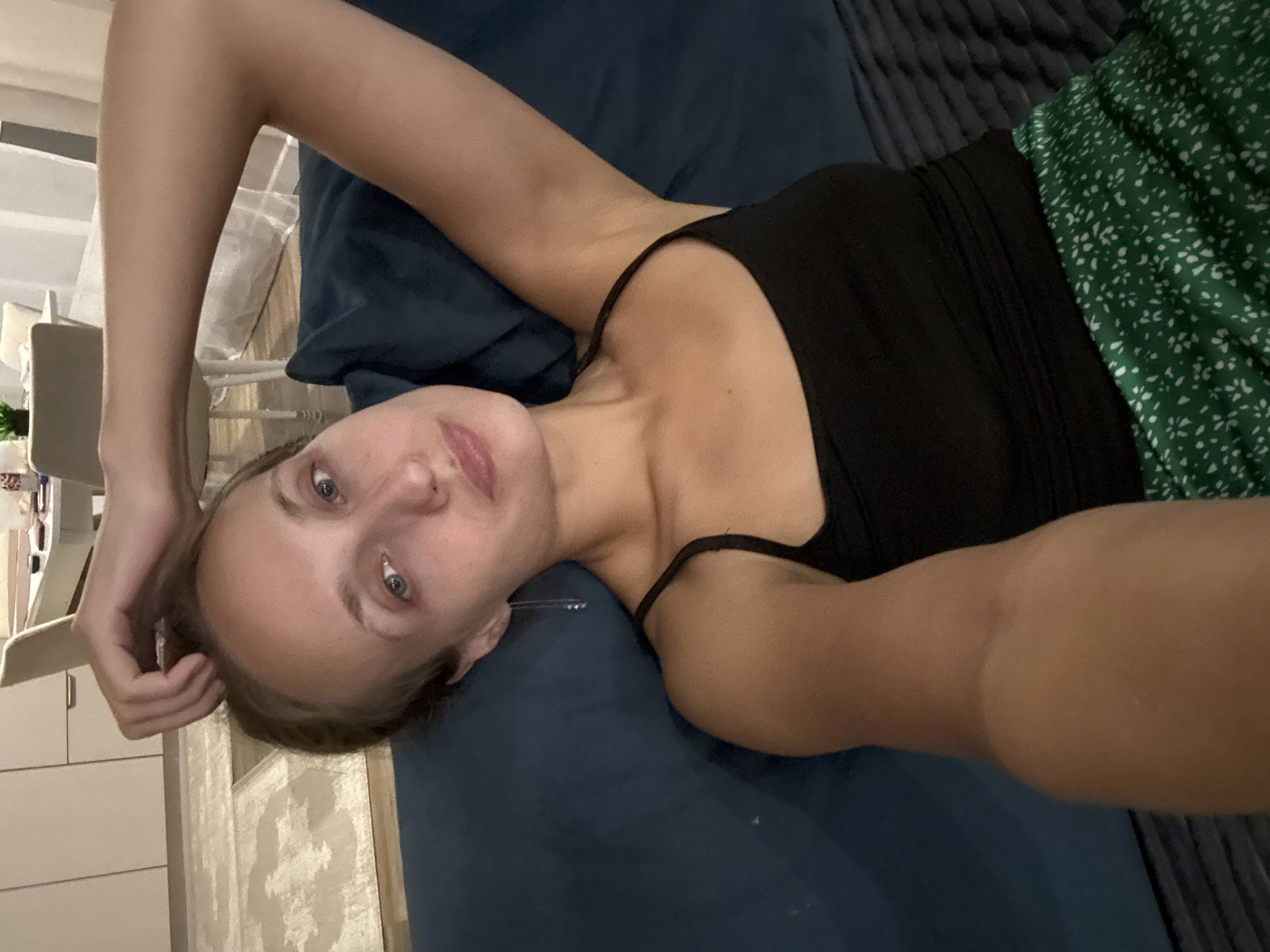 A young woman with light brown hair is lying on a dark blue sofa, with her right arm raised behind her head. She is wearing a black tank top and a green skirt with a white floral pattern. Her eyes are open, and she is looking at the camera with a neutral expression. The background shows a dimly lit room with furniture and what appears to be a dining area. The image is a selfie taken from a high angle. The location of Подшивалово, Russia is not discernible from the image.