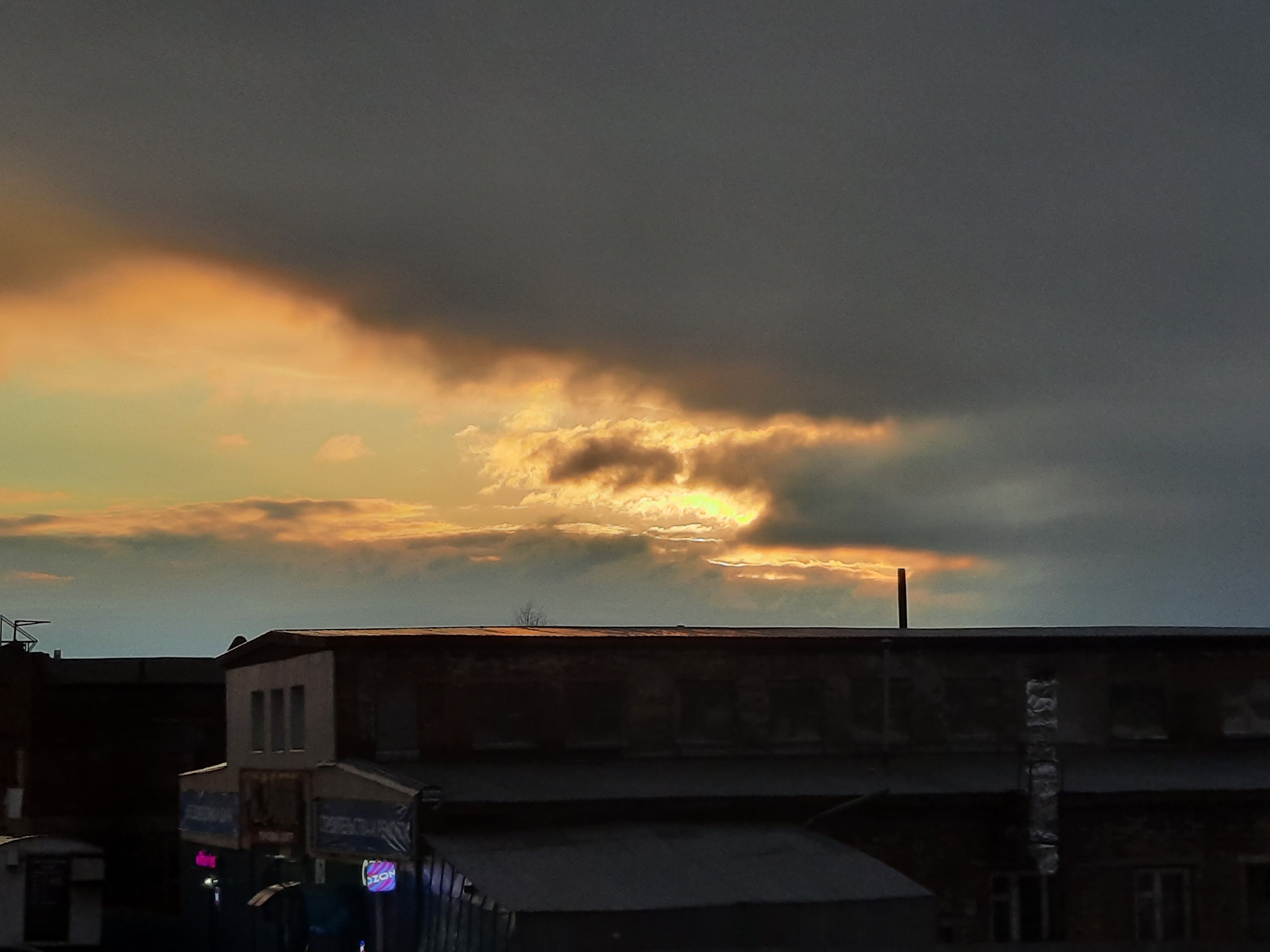 The image depicts a cityscape under a dramatic sky. The location is indicated as Krasnogorskoye, Russia. The sky is predominantly dark grey, but a portion of it reveals an orange-yellow light, suggesting a sunset or sunrise. Clouds are visible, some darker and heavier than others.
In the foreground, there are several buildings, predominantly dark and appearing somewhat aged. There are signs with neon lights visible on the lower level, indicating commercial activity. On the roofs of the buildings, there are a few objects such as a metal pole and aerials. Overall, the image has a muted color palette and a somewhat somber mood.