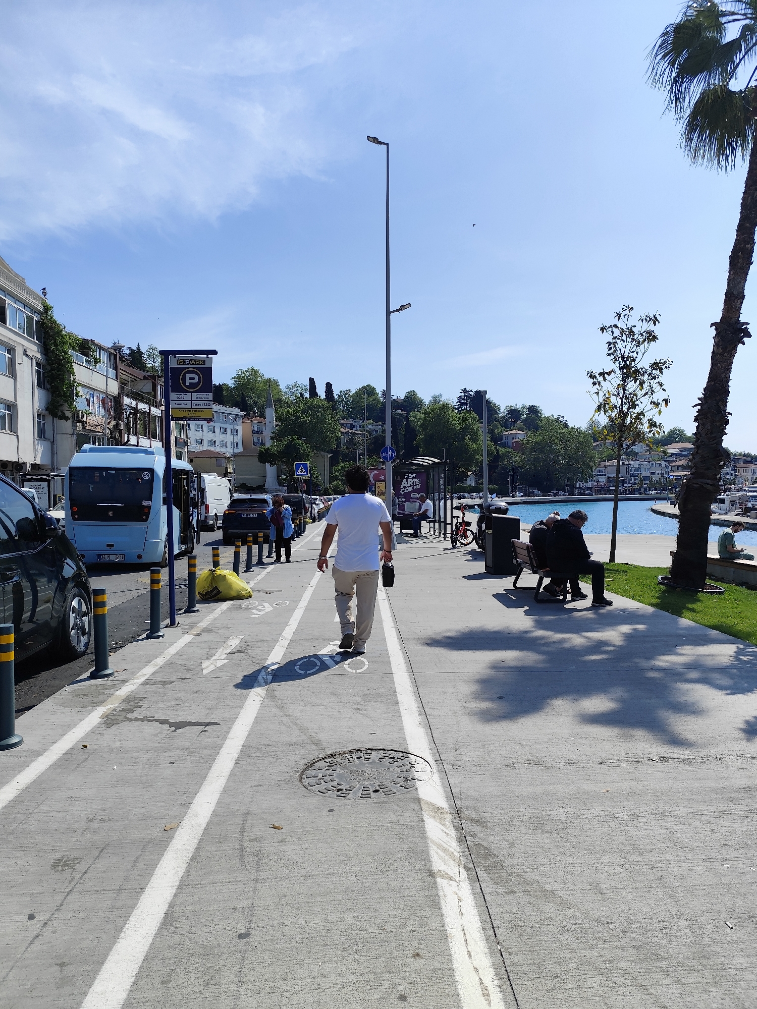 The photo captures a scene in Sarıyer, Turkey, on a sunny day. A man wearing a white shirt and khaki pants walks away from the camera on a concrete path marked with bicycle lane symbols. This path runs parallel to a road with parked and moving vehicles. The man carries a dark object, possibly a bag or bottle, in his right hand. Several people stand near the vehicles and a bus stop sign.
To the right of the path, along the water's edge, are benches where people are sitting. A palm tree stands prominently at the edge of the frame. In the background, across the water, are buildings and trees lining the shore. The water is a vibrant turquoise blue.
The scene includes typical urban elements such as light poles, traffic signs, and a manhole cover on the path. There is a mix of pedestrian and vehicular traffic, typical of a populated city location.