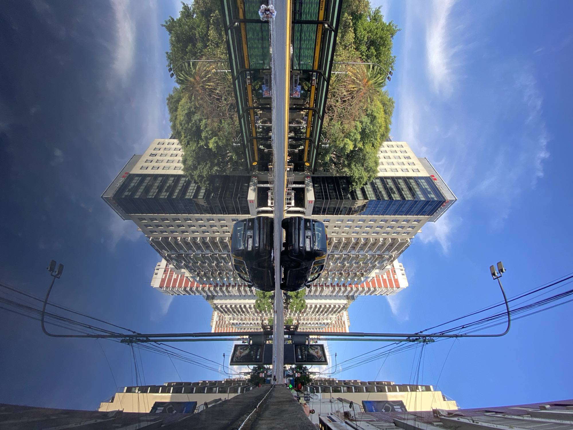 The image shows an inverted view of the Buenos Aires Teleferico cable car in the city of Buenos Aires, Argentina. The view looks upwards, from the bottom station, presenting an upside-down perspective of the upper station, cable car system, and surrounding environment. The building visible in the image is partially a mirrored image of a building, with an architectural style that includes a combination of a modern glass facade and traditional white stone sections with red accents.
The cables of the cable car system are prominent in the lower portion of the image, appearing to stretch downwards towards the viewer. A single passenger is visible in the top of the frame on a cable-car loading platform. There are lush trees that appear mirrored. The sky transitions from a dark blue to a bright blue at the “bottom” of the picture, which suggests the direction of natural lighting. Two cable cars are hanging upside-down in the middle of the image.