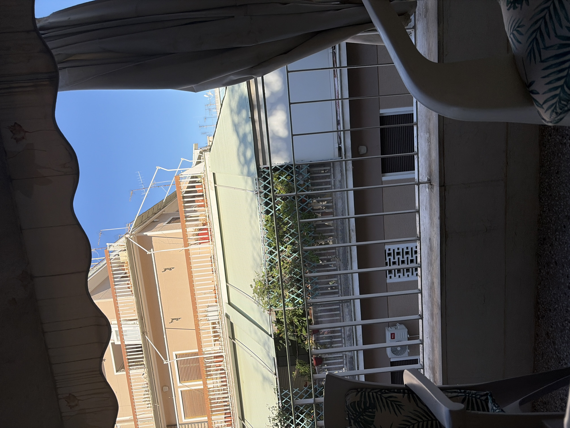 The image captures a view from a balcony in Athens, Greece. In the foreground, parts of white plastic outdoor chairs with a tropical leaf pattern are visible. Beyond the balcony railing, a multi-story building with beige and pale pink facades stands under a clear blue sky. The building features balconies with white railings and wooden accents, some of which have plants in pots. A green retractable awning is extended over one of the lower balconies. In the middle ground, another building facade is visible, with the reflection of clouds on a white surface. An outdoor air conditioning unit with the "TCL" logo is mounted on the wall of this building. The ground beneath the balcony appears to be covered in small stones. The overall scene depicts a residential urban environment on a sunny day.
