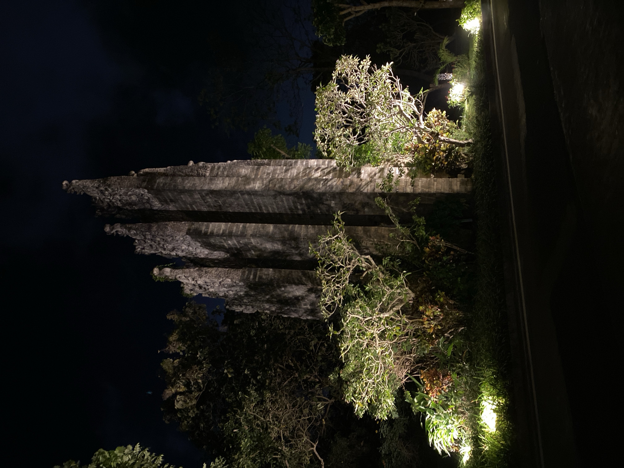 An outdoor, medium shot captures an illuminated ancient stone gate at night. The gate, a towering structure with intricate carvings and multiple spires, dominates the center of the frame. It is made of weathered, grey stone, with vertical lines suggesting the texture of the material. The gate is surrounded by lush vegetation, including trees and shrubs, which are artfully lit by strategically placed yellow spotlights. These lights highlight the foliage and create a dramatic contrast with the dark night sky. A paved road runs along the bottom of the image, with a white line marking its edge. The overall scene evokes a sense of mystery and grandeur, with the ancient architecture standing as a silent sentinel in the darkness.
