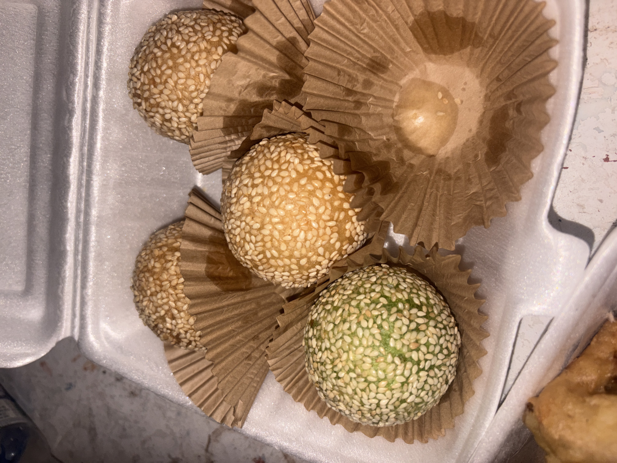 The image displays a close-up, overhead view of four round, sesame-coated pastries or desserts inside a white styrofoam takeout container. Three of the pastries are golden-brown and generously covered in white sesame seeds, while one pastry, located at the bottom right, is distinctly green and also covered in white sesame seeds.
Each pastry rests in its own individual pleated, brown paper liner, similar to a cupcake liner. There is also a fifth empty brown paper liner visible towards the top right, which shows some oil residue at its base, suggesting a pastry may have been removed from it. The styrofoam container has visible textured ridges and edges. A small portion of another golden-brown food item, possibly fried, is visible in the bottom right corner, partially outside the main container.
The lighting appears to be artificial, highlighting the textures of the sesame seeds and the paper liners. There is no visible text in the image. The items are neatly arranged within the takeout container, suggesting they are prepared for consumption or transport.