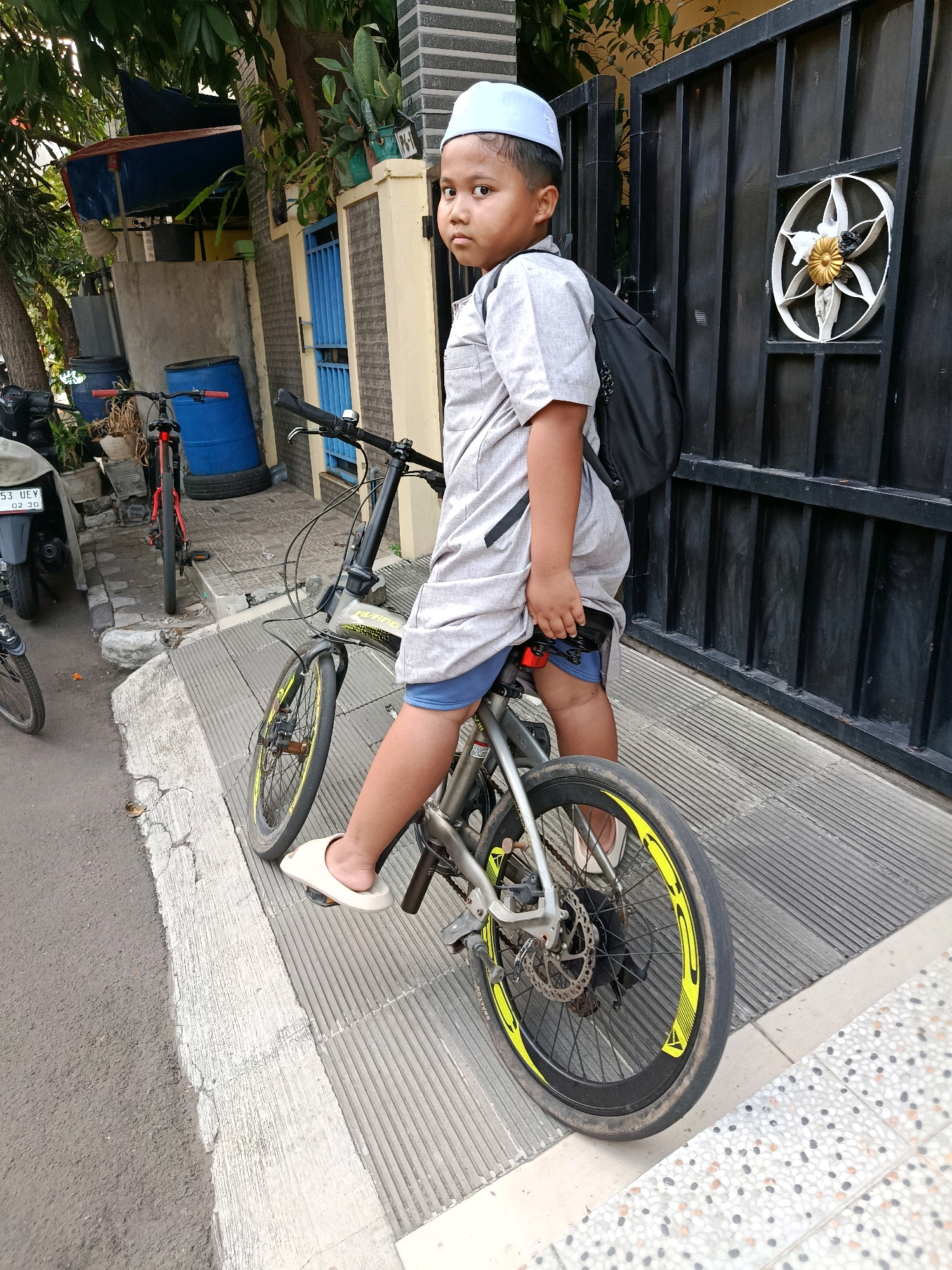 A young boy wearing a white peci cap and a light gray traditional outfit sits on a bicycle in front of a black metal gate. He is positioned on the bicycle from a side-view perspective, with his right leg extended forward. The bicycle has a silver frame and yellow accents on its wheels. The boy is carrying a black backpack. In the background, to the left, there is a blue drum and another red bicycle. Further back, there are some plants and a wall. The image appears to be taken outdoors in an urban or suburban setting, possibly in Bekasi, Indonesia, given the cultural attire and environment.