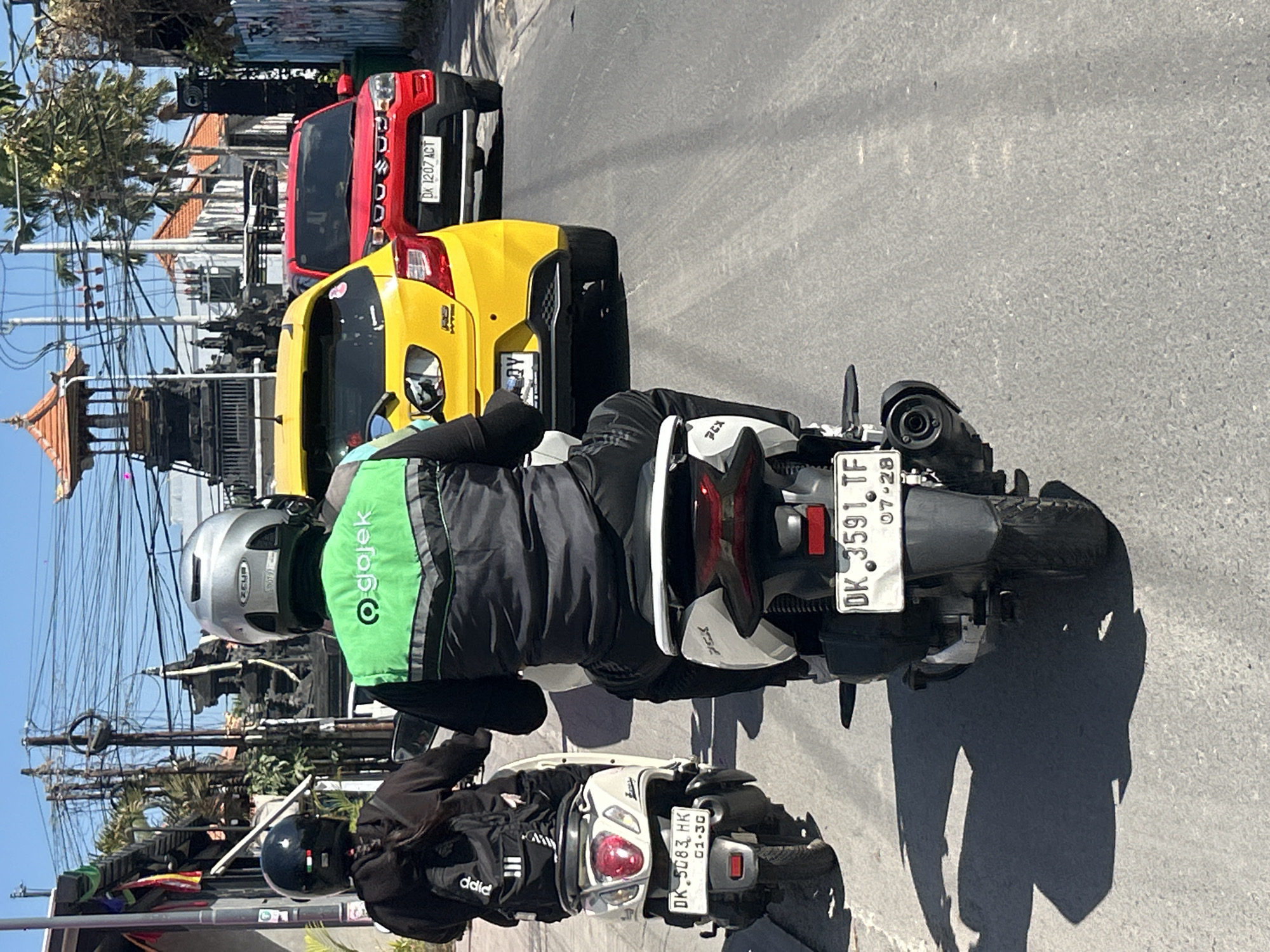 Two individuals on scooters are traveling down a paved street in North Kuta, Indonesia. The scooter in the foreground has a rider wearing a black jacket with a green Gojek vest over it and a silver helmet. The scooter behind them has a rider wearing a black hoodie and a black helmet. The street is lined with buildings and trees, and overhead power lines are visible. In the background, a yellow car and a red car are also on the street. The weather appears to be sunny.