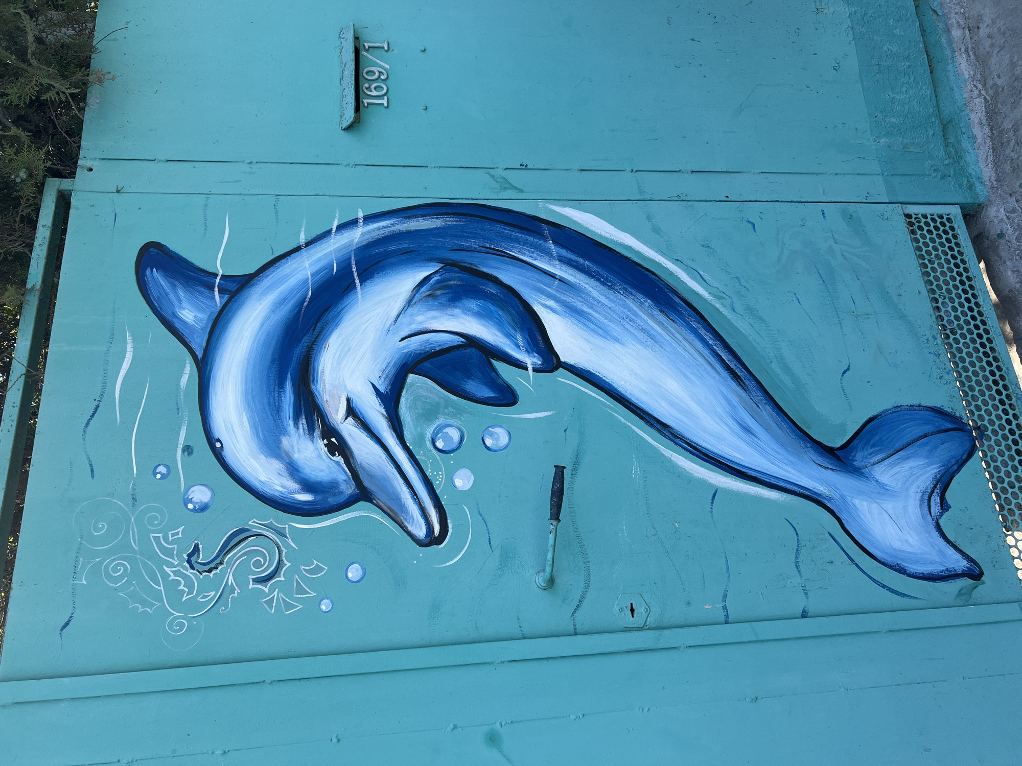 A bright teal metal door is adorned with a large mural of a blue dolphin swimming through stylized waves and bubbles. To the left of the dolphin, a seahorse is depicted in white outline with some blue shading. The dolphin's body is painted in various shades of blue and white, with black outlines defining its form. There are several bubbles of varying sizes scattered around the dolphin, also rendered in white and light blue. A black metal handle is visible on the door, positioned below and slightly to the left of the dolphin's head. To the right of the mural, a small metal mailbox with the numbers "169/1" is attached to the door. The bottom of the door features a metal grate. The mural is painted on the exterior of what appears to be a garage or service building. This artwork is located on the wall of a building at 169 Podgornaya Street in Yeysk, Russia, adjacent to the Yeysk Sea Port. The surrounding environment is not clearly visible, but the presence of a building and a street suggests an urban or semi-urban setting.
