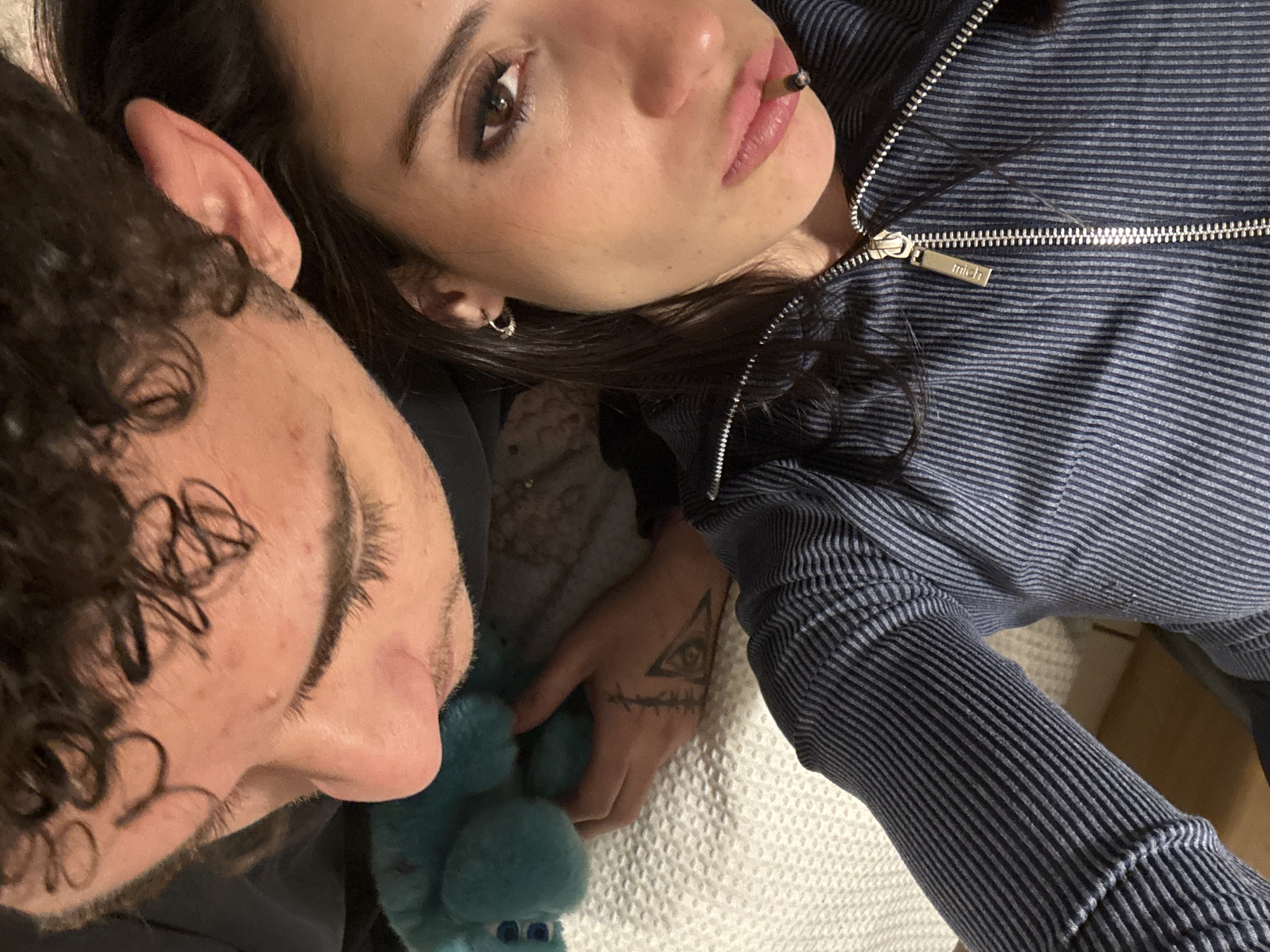 A selfie taken from a high angle shows two people indoors. The person on the right is facing upward, with dark hair, brown eyes, and wearing a striped blue-gray zippered jacket. A small, silver hoop earring is visible in their left ear. They have a tattoo of a triangle with an eye inside it, and a series of short lines below it, on their right hand. They have what appears to be a rolled cigarette in their lips. The person on the left, presumed to be the photographer, has curly hair and is facing downwards, with their cheek near the other person’s hand. A blue stuffed animal sits near the first person. The location is not discernable.