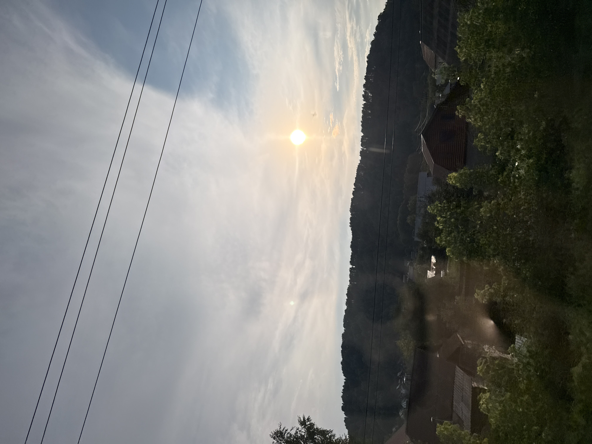 The image depicts a rural landscape under a cloudy sky with the sun visible and casting a bright glow. In the foreground, there are houses and trees, suggesting a residential area. The background features a forested hillside. Power lines are visible stretching across the sky. The scene appears to be captured from a slightly elevated perspective. While the image does not provide specific identifiers, the description of a rural setting with houses and forested hills is consistent with areas around Gorno-Altaysk, Russia. There are no people or animals visible in the frame, and no discernible actions or interactions are taking place. The overall impression is a serene, natural environment with human habitation.