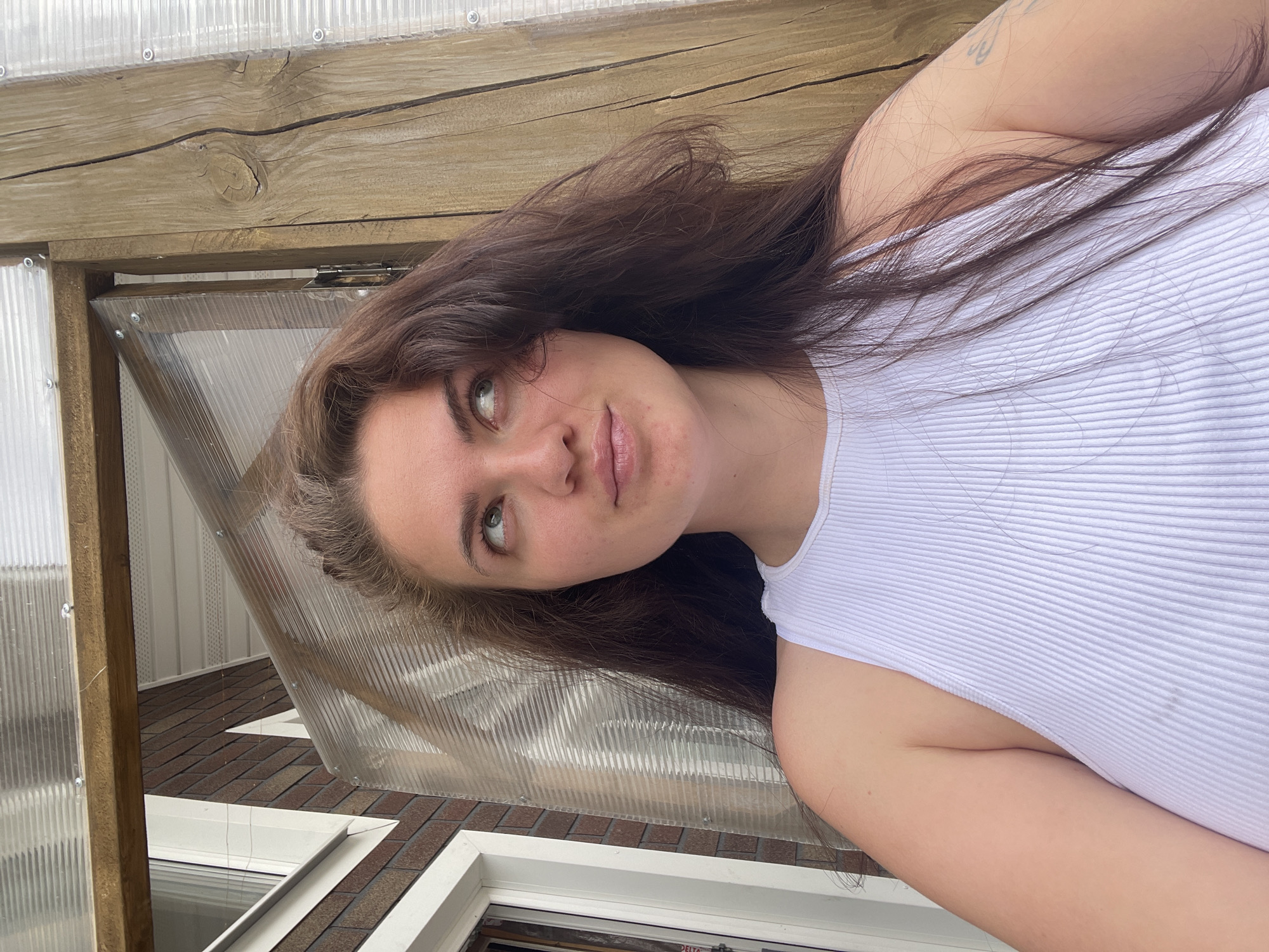 A young woman with long, dark brown hair and fair skin is pictured indoors, seemingly in a greenhouse or enclosed porch-like structure. She is wearing a white ribbed tank top. The woman's face is angled to her left, with her eyes looking slightly upwards. The background includes translucent corrugated plastic panels attached to a wooden frame. Below, portions of a window and brickwork are visible. The scene is indoors, with natural light present. There is no evidence to confirm that the location is in the city of Мосметаллоконструкция Онт, Russia.