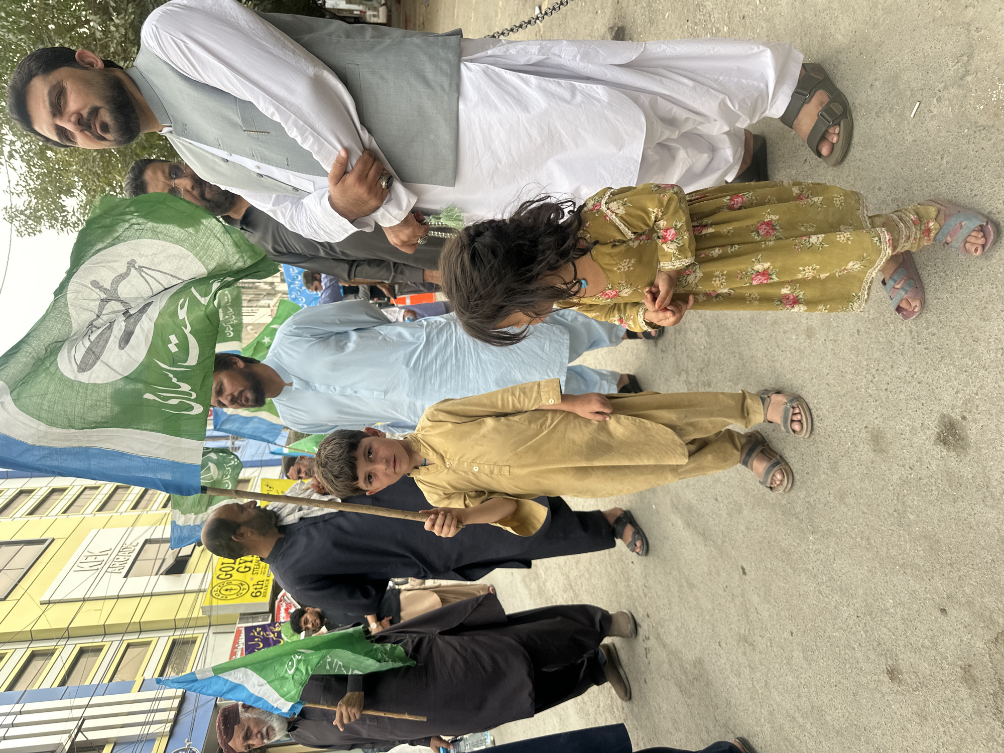 An outdoor, eye-level, medium shot captures a group of people, including two children, standing on a paved surface in Quetta City Tehsil, Pakistan. In the foreground, a young boy, positioned slightly left of center, wears a beige tunic and pants and holds a wooden stick. To his right, a young girl, facing left, is dressed in a floral patterned dress and pink sandals. Behind them, a man in a light blue tunic stands next to a man in a grey vest and white tunic. Several large flags, predominantly green and white with blue accents, are visible, some bearing script. In the background, a yellow building with the sign "KEK ARCADE" is prominent, alongside other structures and trees. The overall scene suggests a gathering or procession.