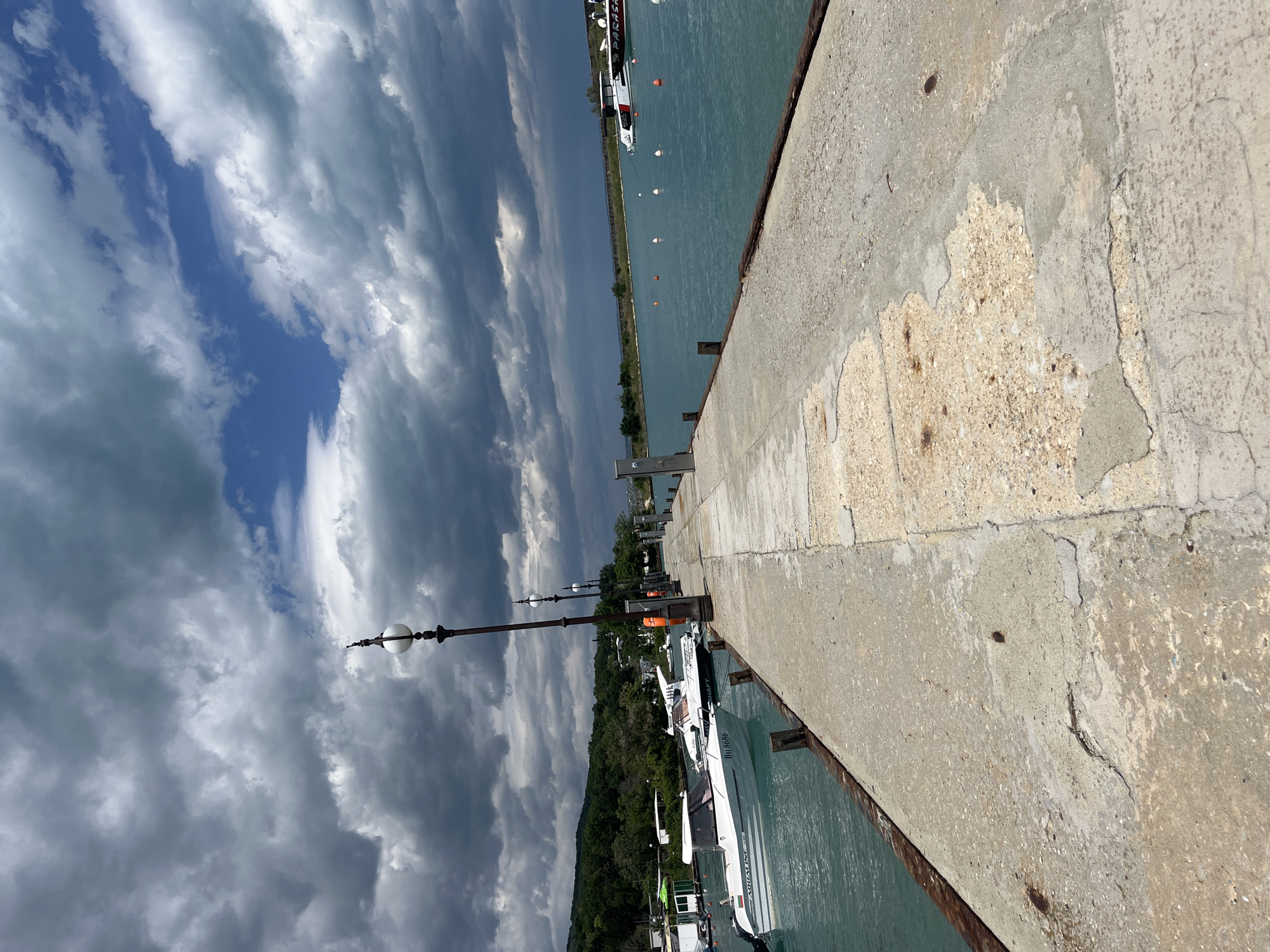 The image shows a concrete pier with a cloudy sky above and water on either side. A row of lampposts lines the pier. Several boats are docked along one side of the pier. The exact location cannot be determined from the image, but it is in the city of Varna, Bulgaria.