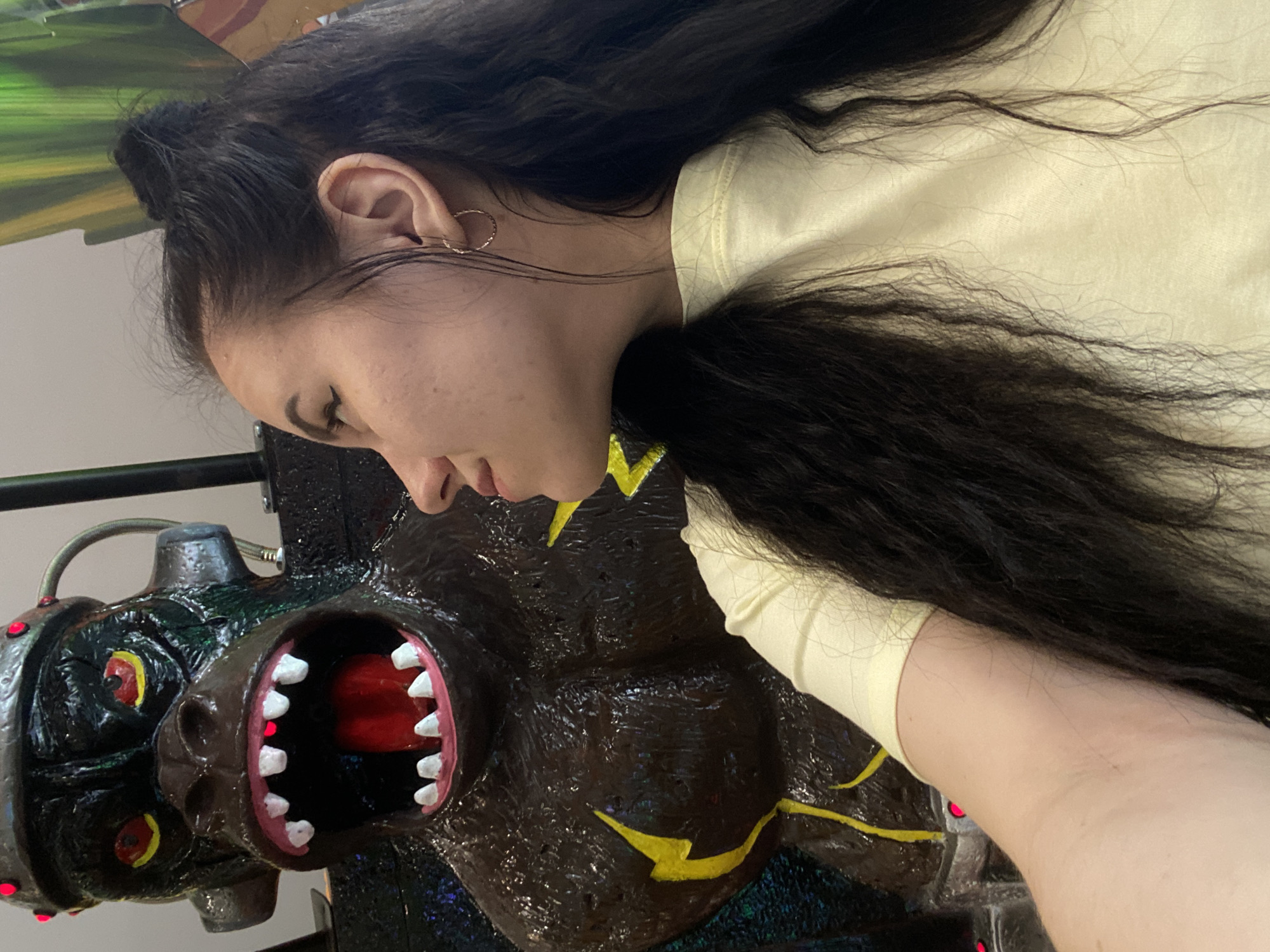 A woman with dark hair pulled into a bun is lying on a dark brown and black mechanical gorilla head with a large open mouth showing red inside and white teeth. The gorilla has yellow lightning bolts painted on its chest. The woman is wearing a light yellow shirt. The location is not definitively identifiable as Perm, Russia based solely on the provided image.