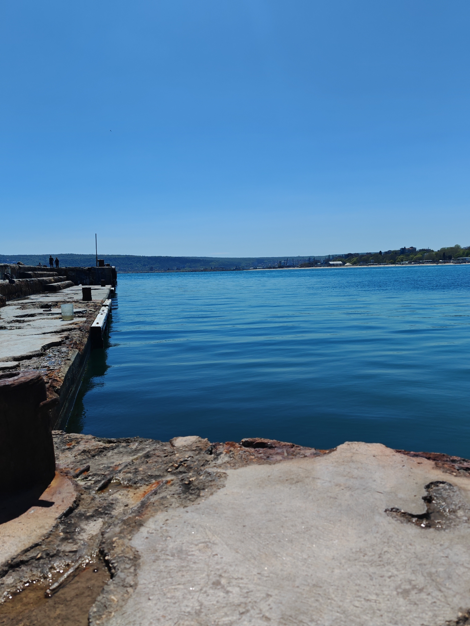 The image captures a seaside view from a concrete pier in Varna, Bulgaria. The clear blue sky meets the calm, blue water of the Black Sea on the horizon. Across the water, a shoreline is visible with a mixture of trees, buildings, and industrial structures. On the pier, weathered concrete structures are interspersed with metal fixtures, showing signs of rust and decay. In the distance, two figures stand on the pier, near a white cup.