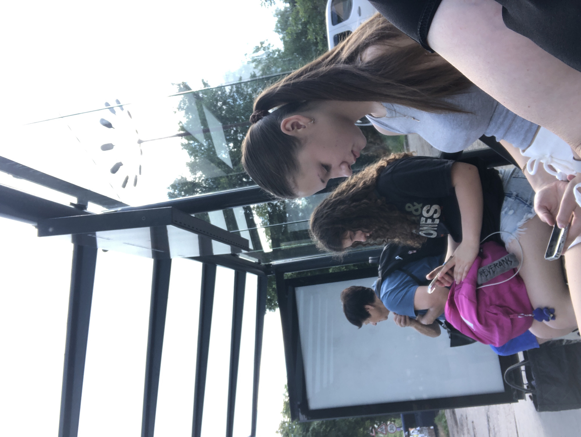 The image shows a group of young people at a bus stop, likely in Chernihiv, Ukraine. A teenage girl with her hair in a ponytail is wearing a grey tank top and white shorts, looking down at her phone. To her left, another young woman with curly hair is wearing a black t-shirt and denim shorts, also engrossed in her phone. In the background, an older woman in a blue shirt and dark pants is seated, looking to her side. She is wearing a backpack. A bright pink backpack with a blue keychain is visible in the foreground. The bus stop has a metal frame and glass panels, with a billboard on the left side. Trees and a white vehicle are visible in the background through the glass. The sky is bright and overcast.