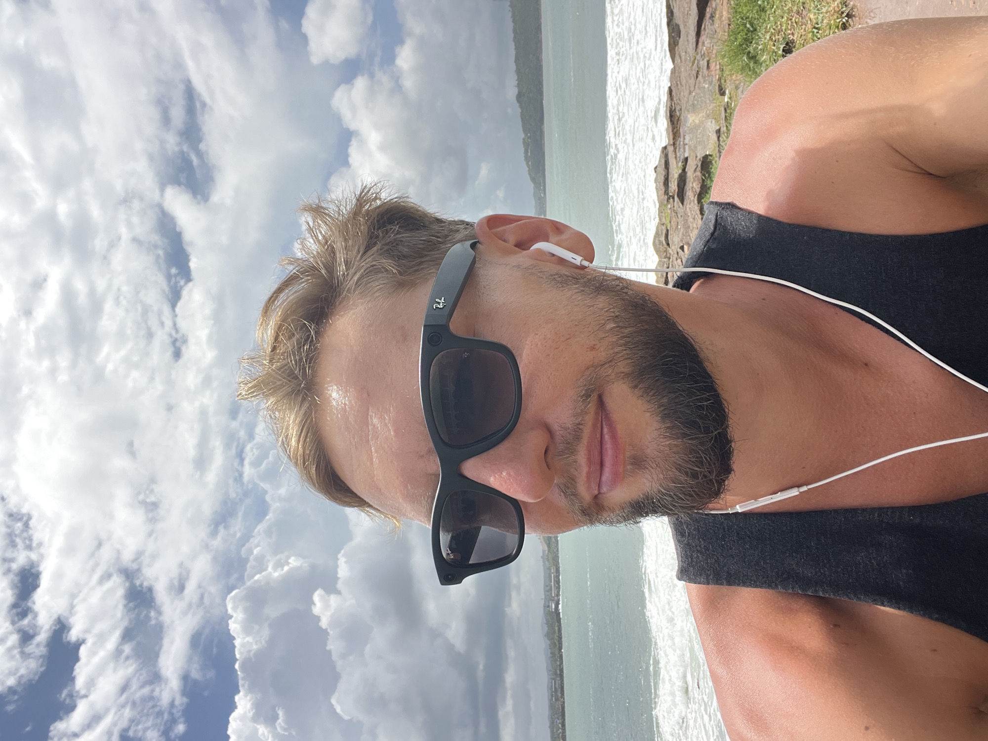 A man with blonde hair and a beard, wearing black sunglasses and white earbuds, takes a selfie in Galle, Sri Lanka. He is wearing a black tank top and is outdoors with a cloudy sky and the ocean behind him. The ocean has waves and a rocky shoreline.