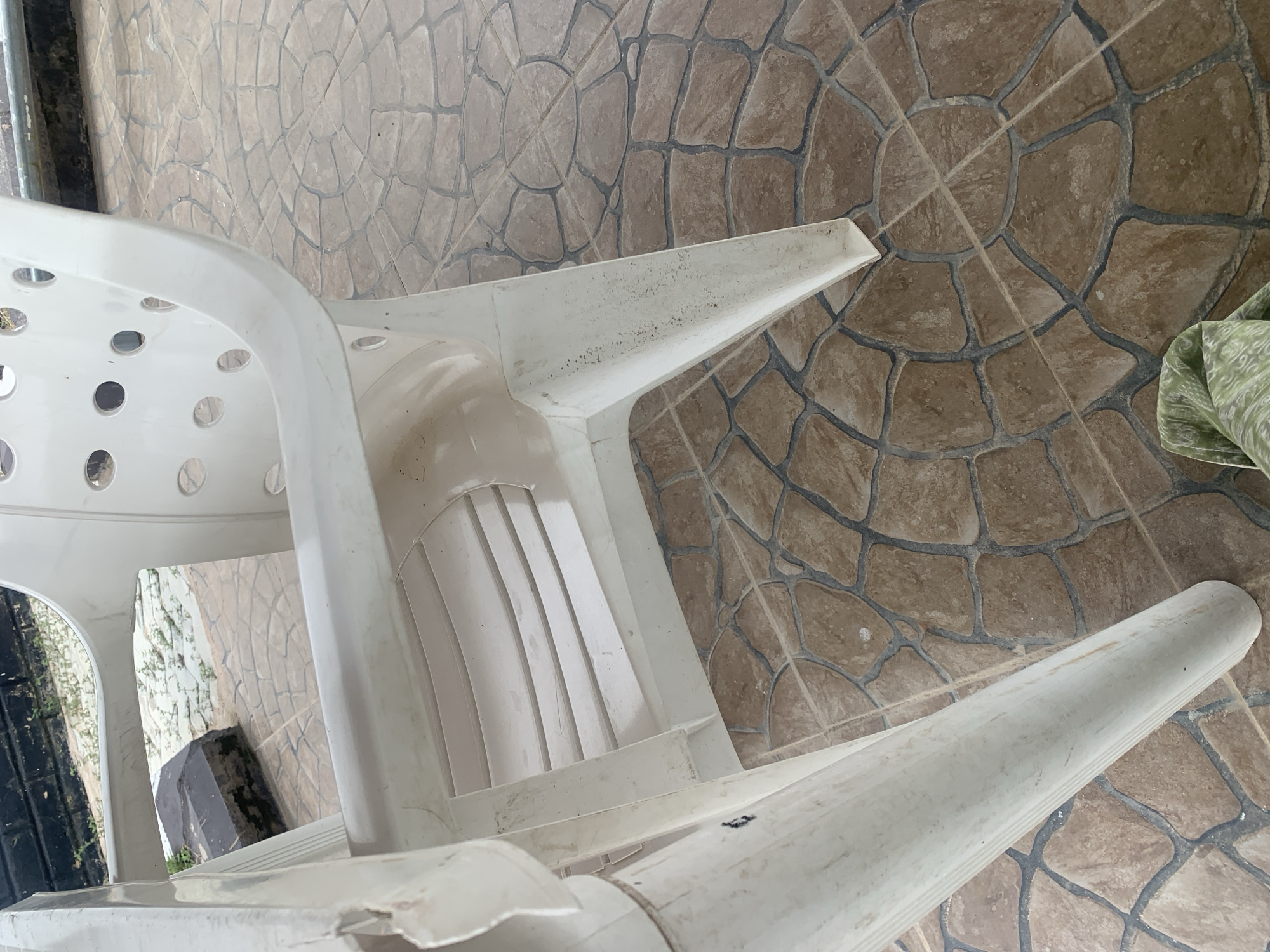 The image shows a close-up of a white plastic chair standing on a patterned tiled floor. The chair appears to be outdoors, with some vegetation and a dark structure visible in the background through the chair. The chair has a damaged section on its armrest and shows signs of wear and dirt. The floor tiles are a mix of brown and tan with dark grout lines arranged in a circular pattern. A green and white patterned fabric is visible in the lower right corner of the image. The scene is consistent with an outdoor seating area in Jalingo, Nigeria.
