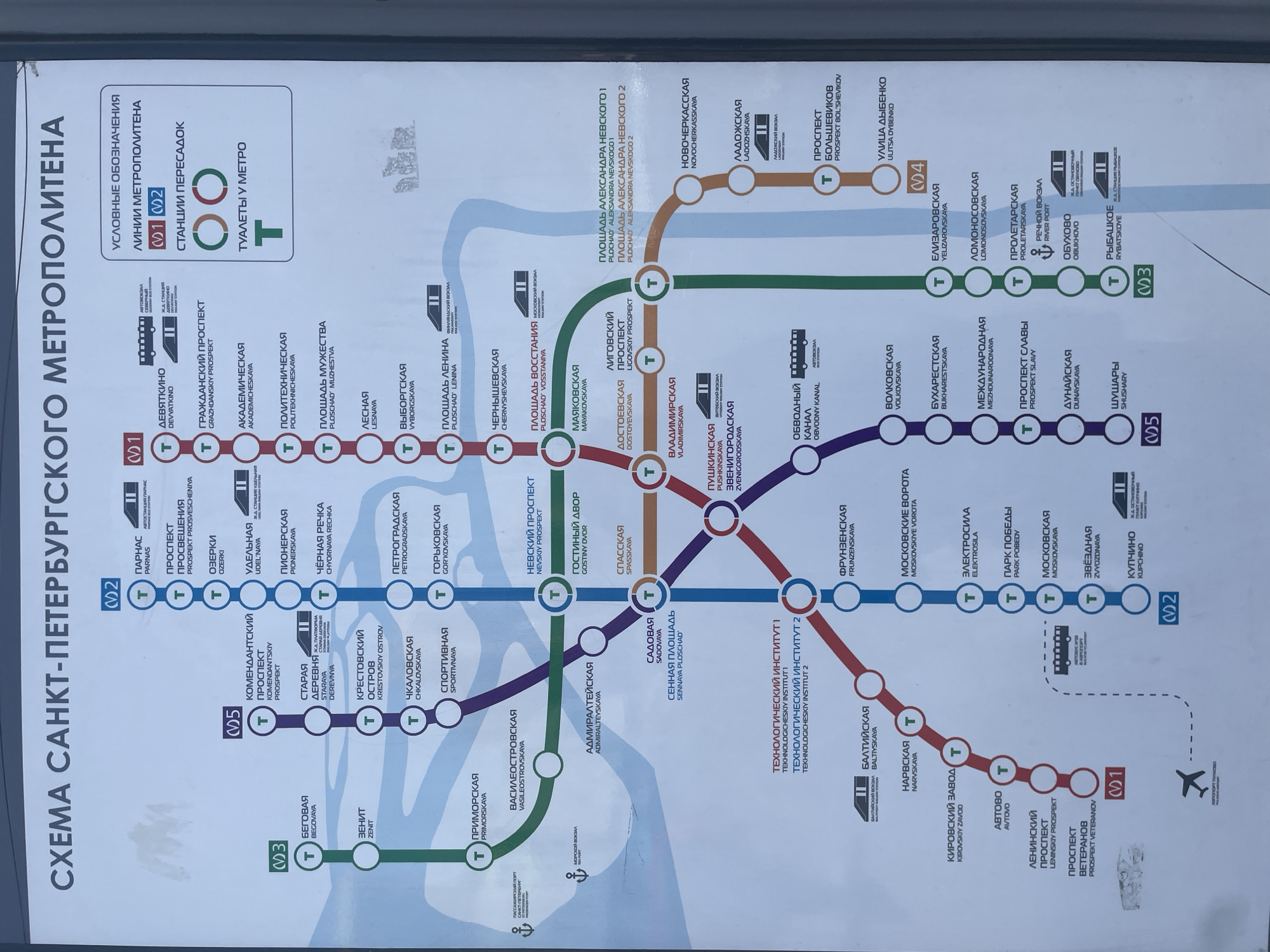 The image is a photograph of a diagram of the Saint Petersburg Metro system in Russia. The diagram displays various metro lines, denoted by different colors and numbers (1, 2, 3, 4, 5), and the stations along these lines. The station names are provided in both Russian and English. Key transfer stations are highlighted. A legend in the upper right corner explains the meaning of symbols used on the map, including metro line identification, transfer stations, and the presence of toilets. The overall presentation is clear and functional, designed for public information.