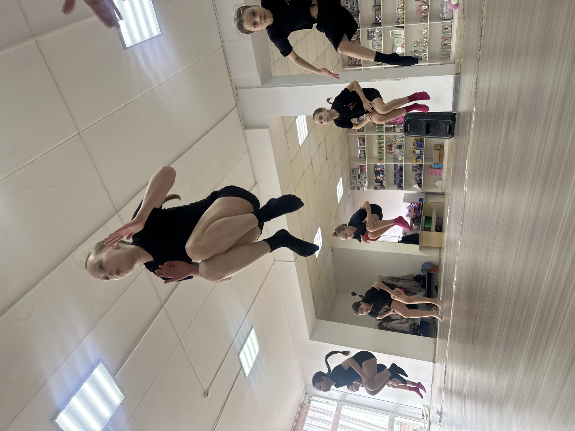 A group of female dancers are practicing in a dance studio in Votkinsk, Russia. The studio has a mirrored wall that reflects the dancers. The dancers are wearing black leotards and socks. Some of the dancers have pink socks. There are shelves with trophies and photos in the background. The floor is made of wood. The ceiling has fluorescent lights.