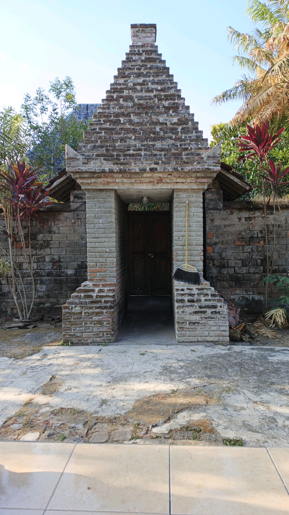 The image shows an entranceway to a building constructed of brick and stone. The entrance is framed by a weathered brick structure with a stepped, triangular facade. To the left and right of the entrance are walls constructed of similar brick and stone. Red-leafed plants are visible on either side of the entrance, and palm fronds are visible in the upper right corner. A broom with a yellow handle leans against the right side of the entrance. The ground in front of the entrance is a cracked concrete patio. The background reveals a blue sky and greenery. This scene is located in Wajak Kidul, Indonesia.