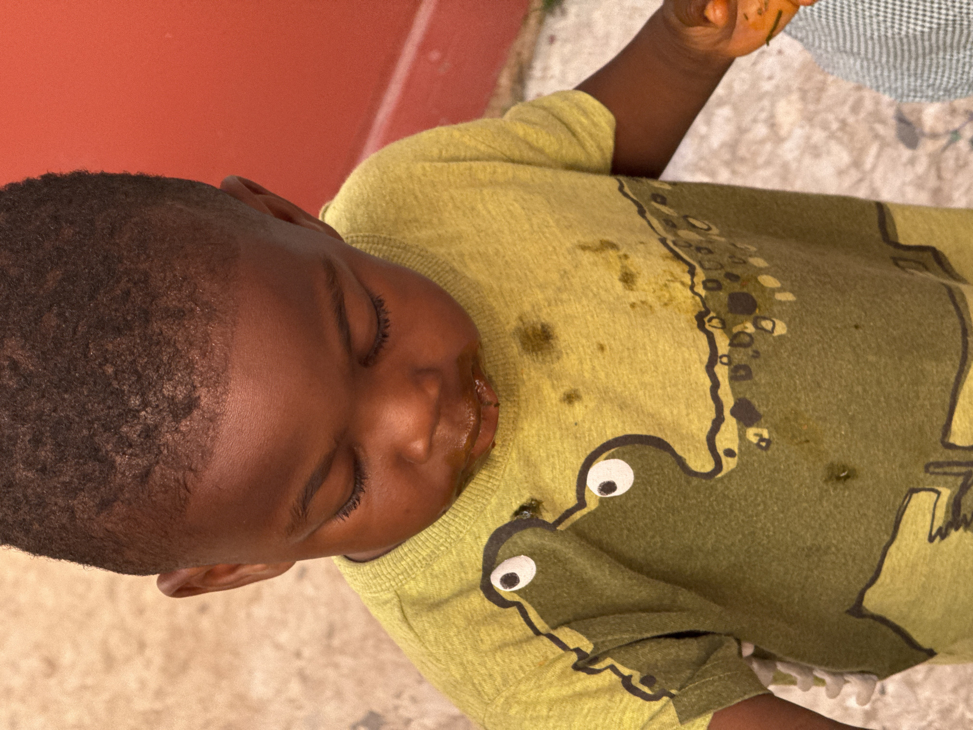 A young Black child with short, dark hair and closed eyes is shown from an overhead angle. The child is wearing a yellow-green t-shirt with a large, stylized crocodile or alligator graphic on the front. The shirt has visible food stains. The background includes a portion of a red surface and a light-colored concrete or tiled ground. There is no evidence to confirm that this photo was taken in Abuja, Nigeria.
