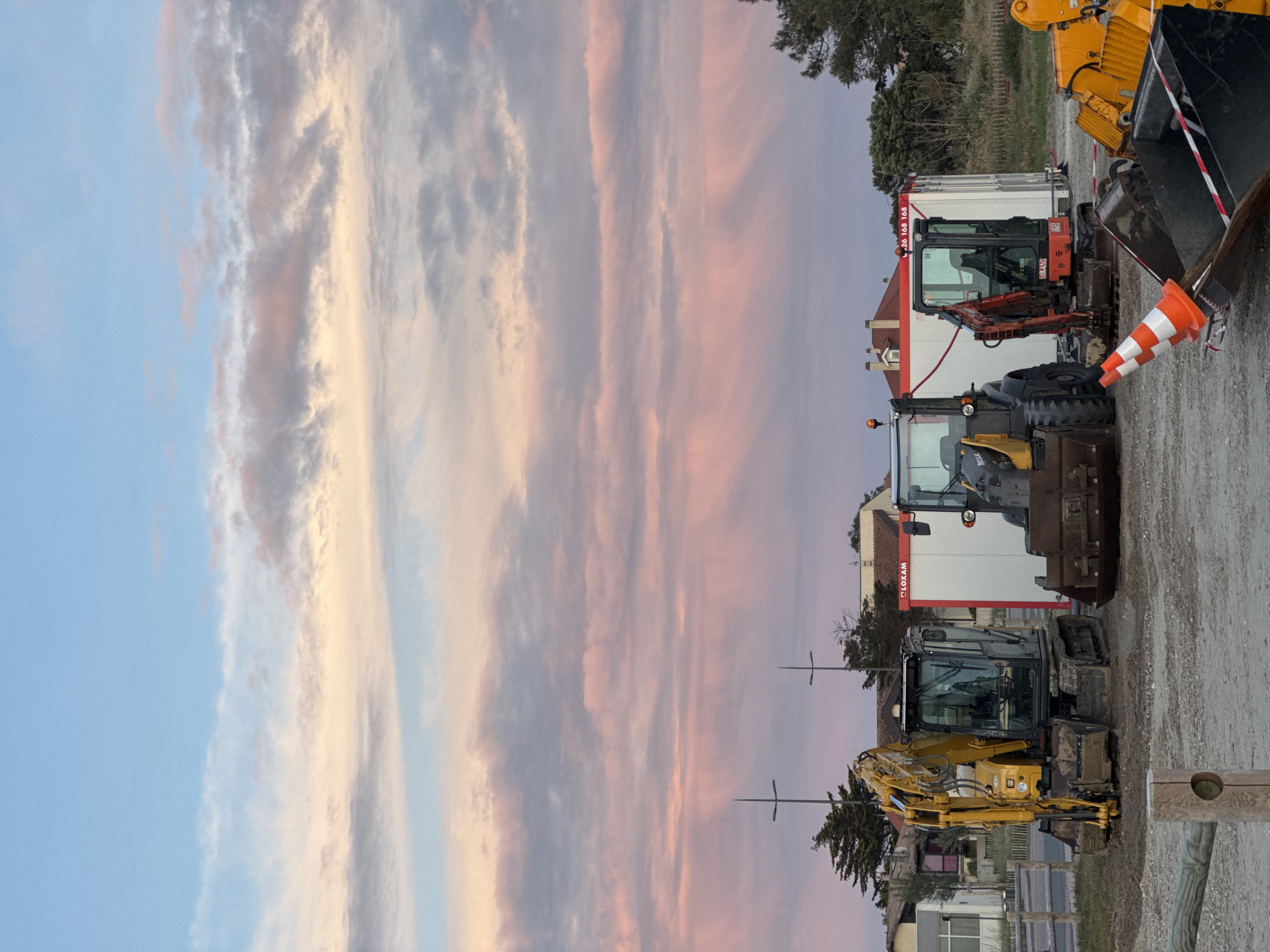 The image shows an outdoor scene in Soulac-sur-Mer, France at dusk. The left side of the image is dominated by a colorful sky with streaks of blue, pink, and grey clouds. The right side features several pieces of heavy construction equipment parked on a gravelly surface. These include excavators and other construction vehicles. A row of low-lying buildings is visible behind the construction equipment. An orange and white traffic cone is also visible.