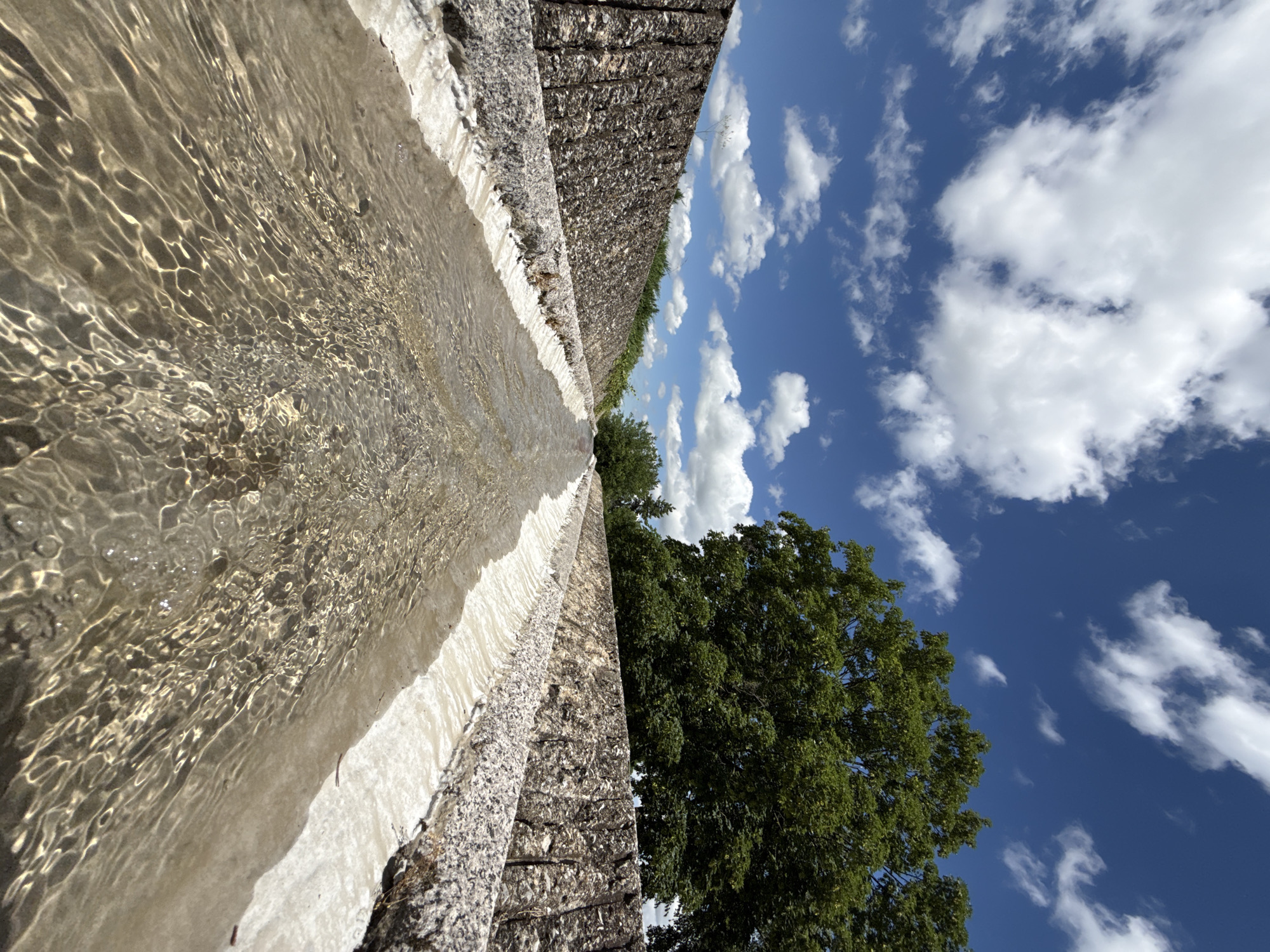 The image depicts a man-made water channel with clear water flowing through it. The channel is flanked by stone walls that are constructed from small, irregularly shaped stones. The water is shallow and exhibits rippling patterns on its surface, reflecting the light from the sky. The background features lush green trees and a bright blue sky filled with fluffy white clouds. The perspective suggests the photo was taken from a low angle, looking down the length of the water channel. This water channel is part of the "Water Cascade" in Shumen, Bulgaria, a prominent feature of the city's landscape.