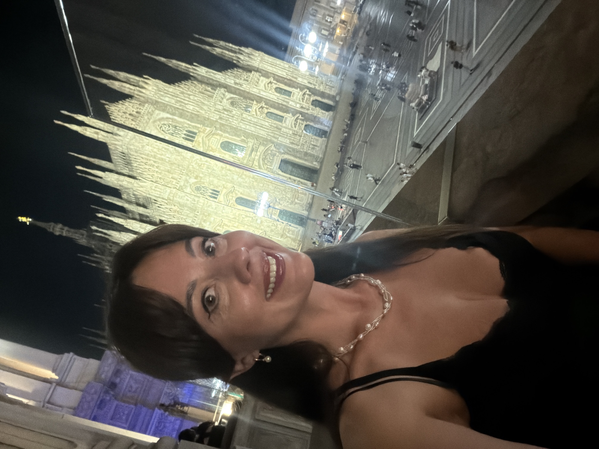 An exterior, eye-level, night-time selfie is captured from a slightly elevated vantage point, looking down onto a public square. The photographer, a woman with dark hair and bangs, is positioned in the foreground, smiling broadly. She is wearing a black spaghetti-strap top with lace detailing along the décolletage and a multi-strand pearl necklace. A single pearl earring is visible on her left ear.
In the background, the Duomo di Milano, a magnificent Gothic cathedral, is illuminated by artificial lights. The ornate facade and spires of the cathedral are clearly visible. The square in front of the Duomo is paved and populated by a moderate number of people, some of whom are walking, while others are seated or gathered in small groups. Streetlights and building lights further illuminate the scene. The overall impression is one of a vibrant evening in Milan, Italy.