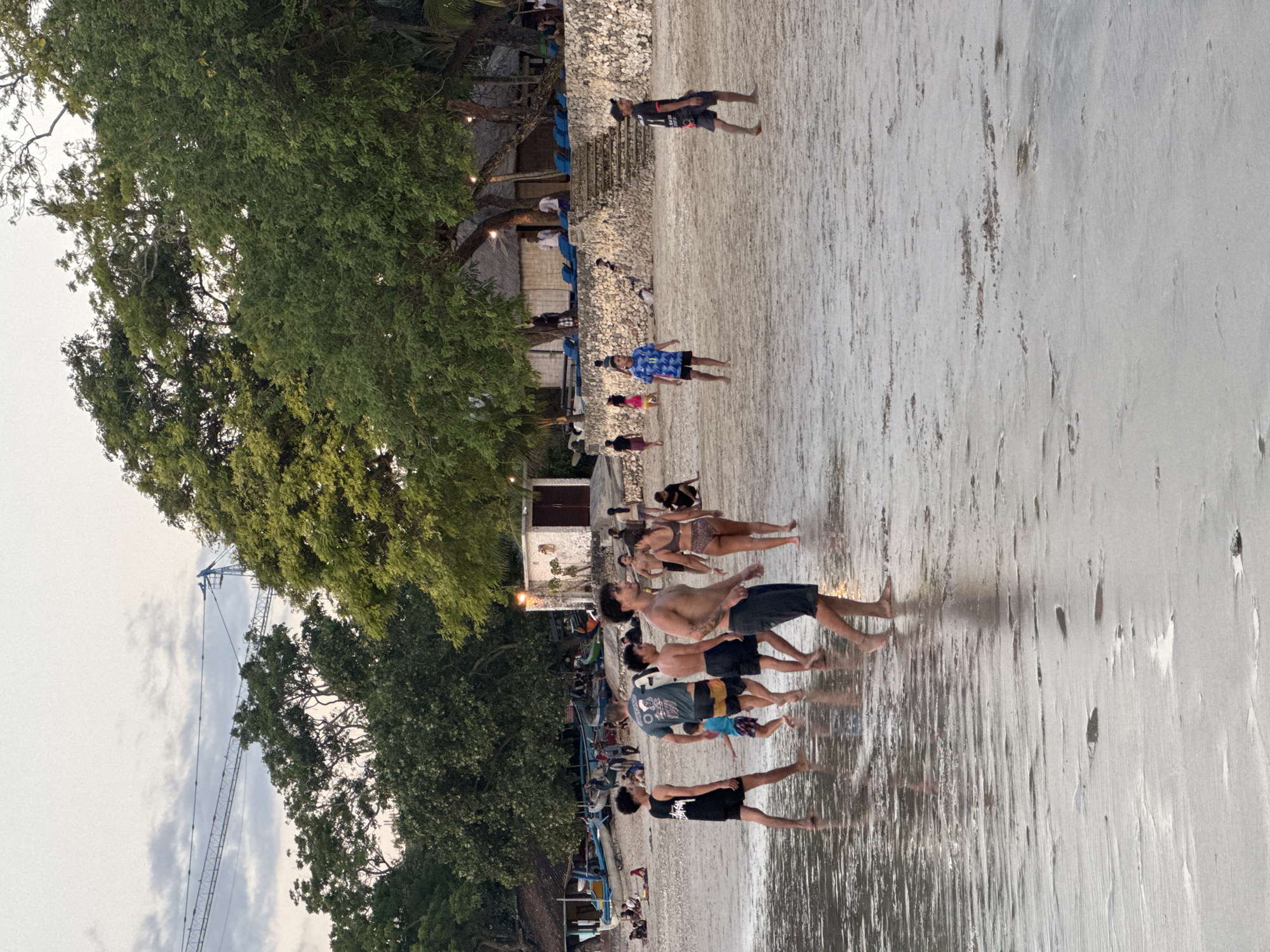 The image depicts a beach scene at dusk in Kuta Selatan, Indonesia. A group of people are standing on the wet sand, some appearing to be engaged in a synchronized activity, possibly a form of exercise or dance, as they are all facing the same direction and in similar poses. The lighting suggests it is either early morning or late evening, with the sun low in the sky.
The background reveals a shoreline with scattered boats, suggesting it's a populated area. Further inland, there are structures that look like beachfront buildings, some with thatched roofs, and trees that add to the tropical ambiance. The sky is overcast, and the overall mood of the scene appears relaxed and communal. The wet sand reflects the dim light, creating a glossy effect. No visible text is present in the image.